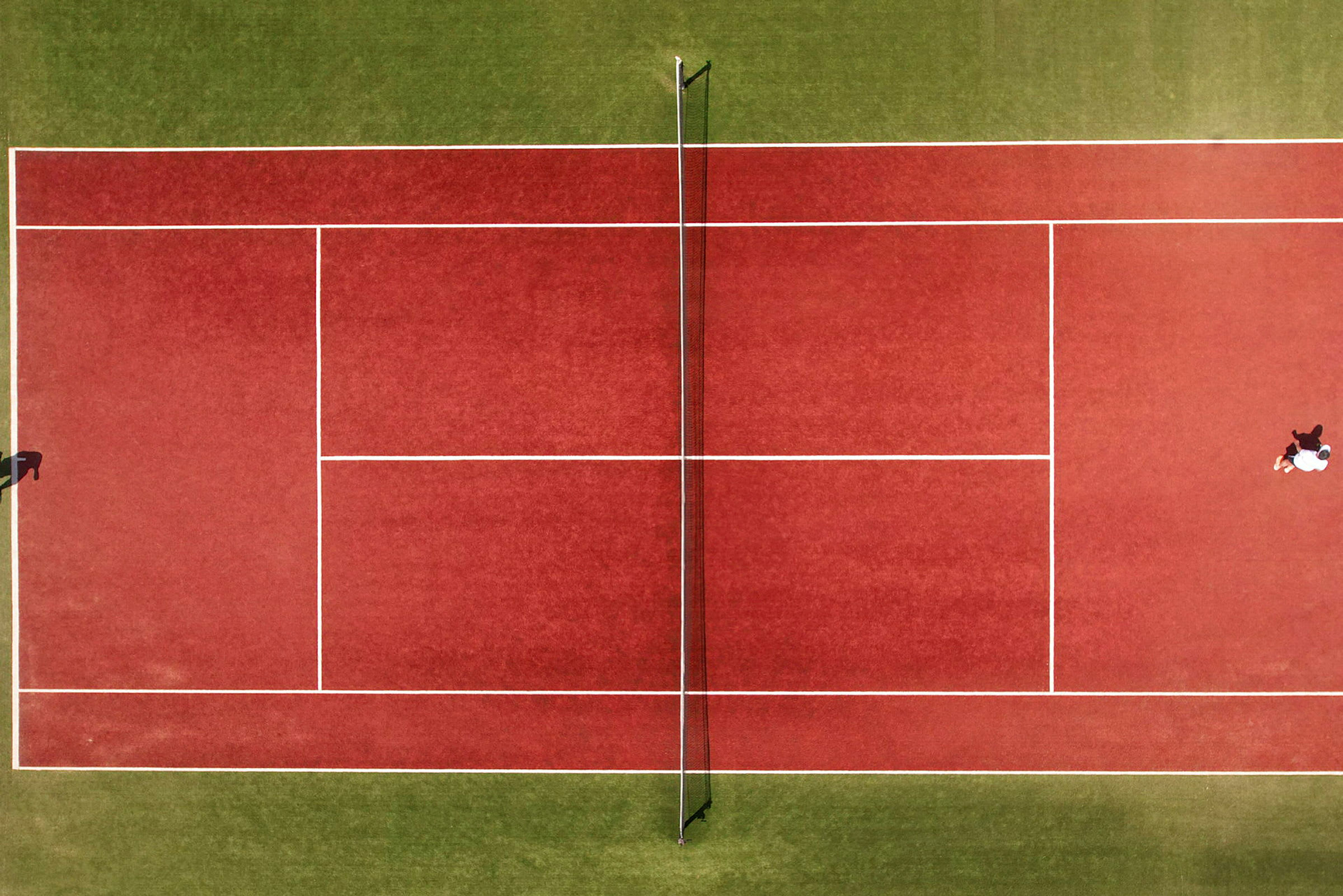 Flyfoto av en rød tennisbane med nett, to personer spiller. (AI-generert tekst)