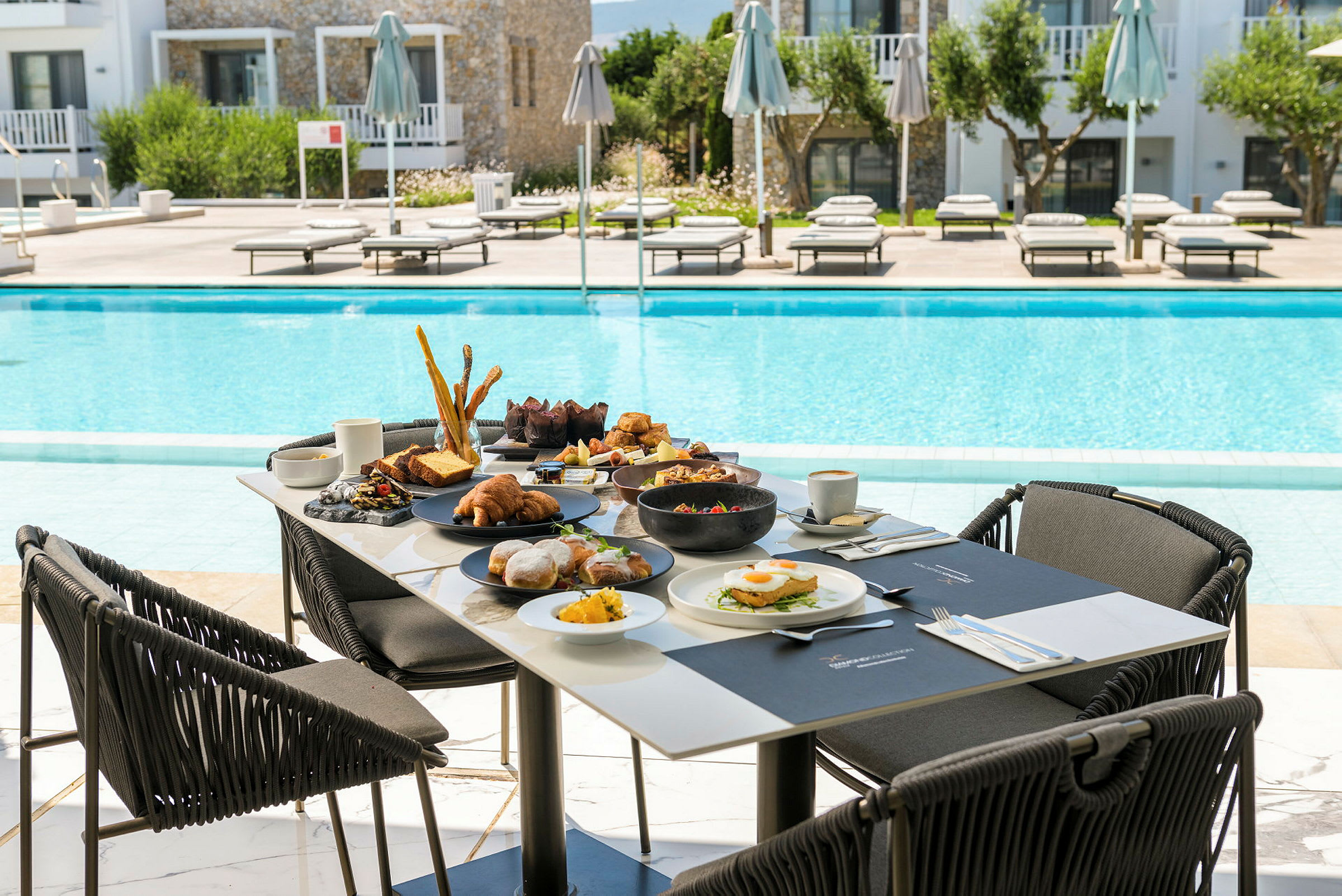 Frokostbord ved et basseng med en resort i bakgrunnen. (AI-generert tekst)