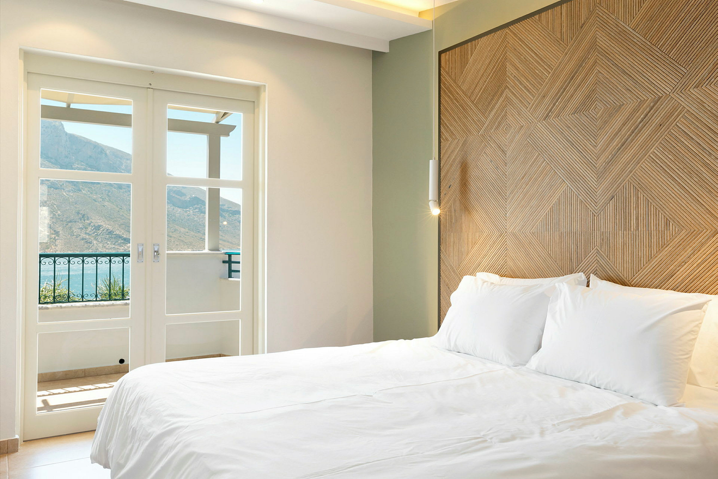 Et hotellrom med en stor hvit seng, et dekorativt tremønstret hodegjerde og glassdører som åpner mot en balkong med fjell i bakgrunnen. (AI-generert tekst)
