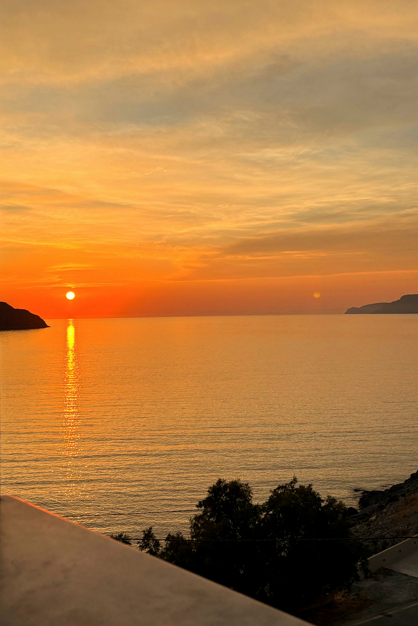 En solnedgang over havet, med oransje og gule farger på himmelen, noen trær i forgrunnen og en klippe til venstre. (AI-generert tekst)