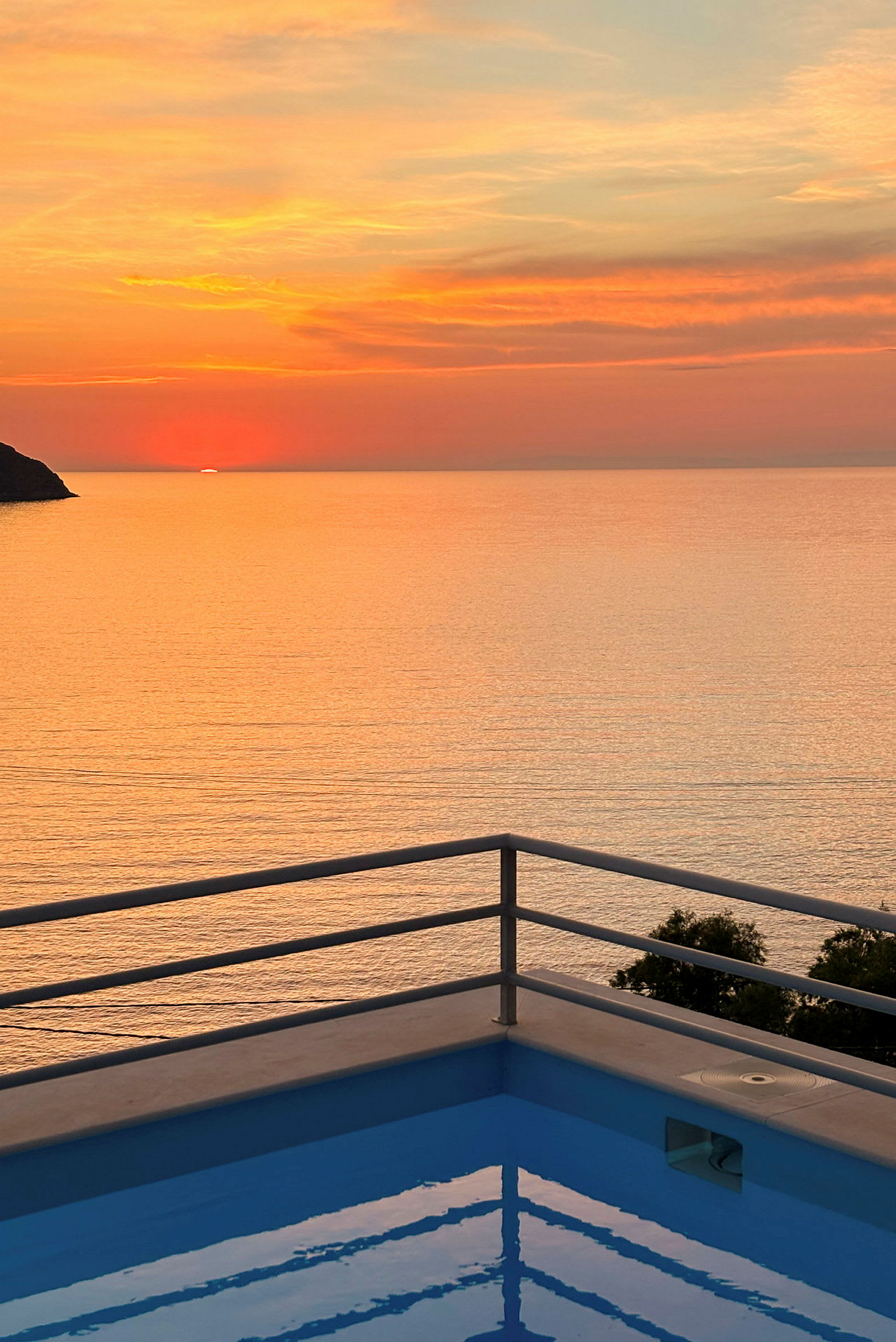Solnedgang over havet med et basseng kantet med et rekkverk i forgrunnen. (AI-generert tekst)