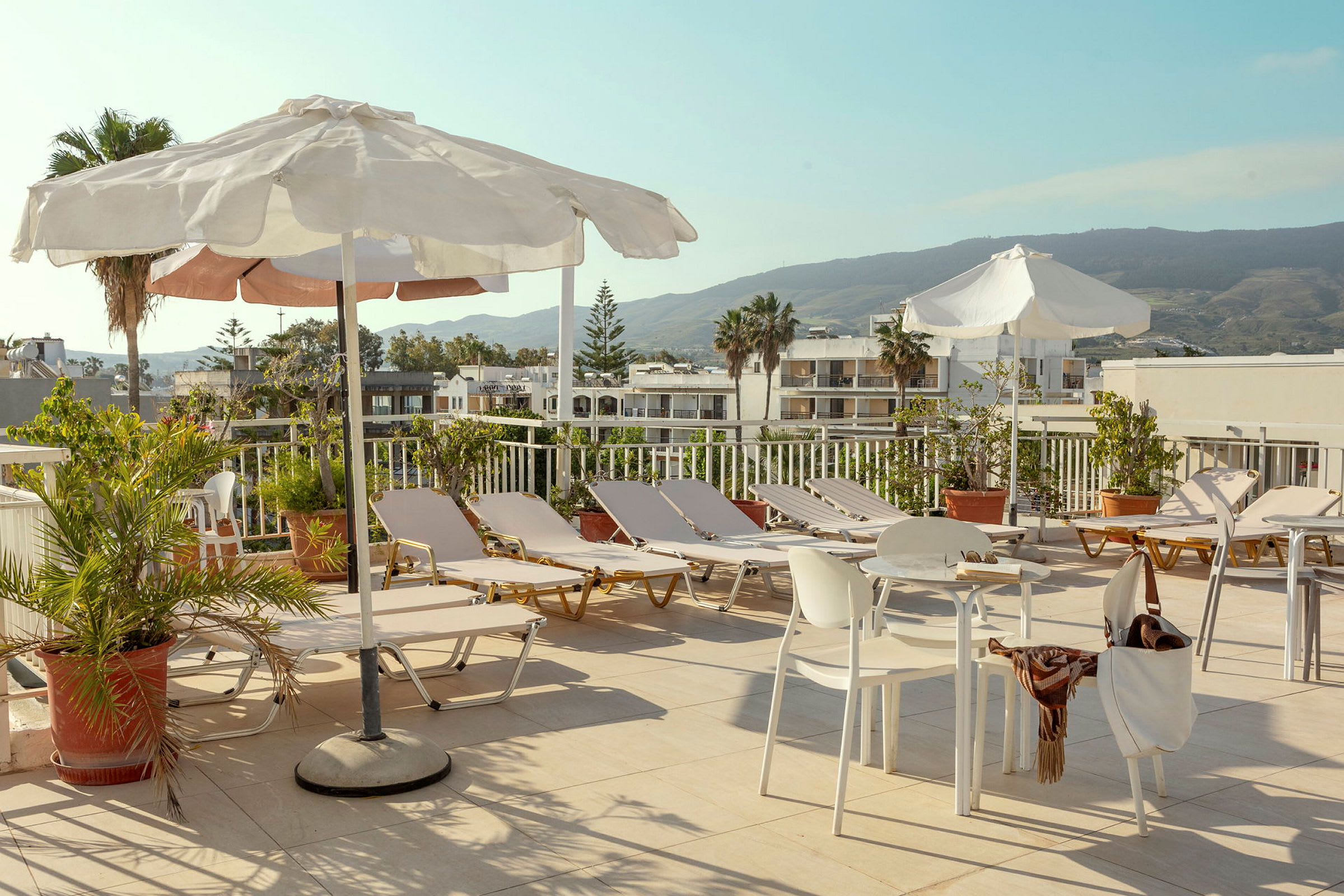 En solterrasse med solsenger, parasoller og bord samt stoler omgitt av grøntområder med utsikt mot bygninger og fjell. (AI-generert tekst)