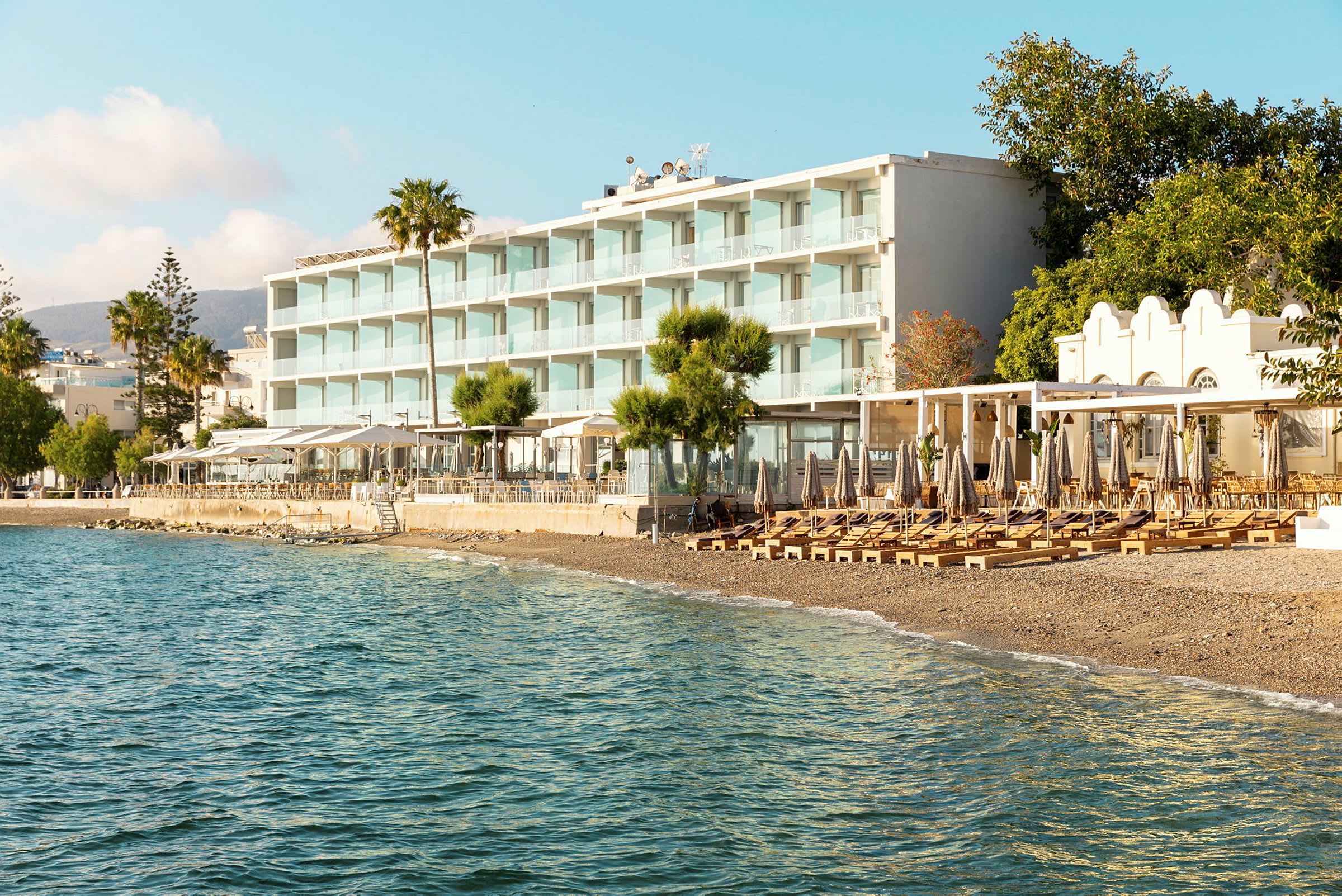 Utsikt mot et strandhotell med solsenger og parasoller på stranden og havet i forgrunnen. (AI-generert tekst)