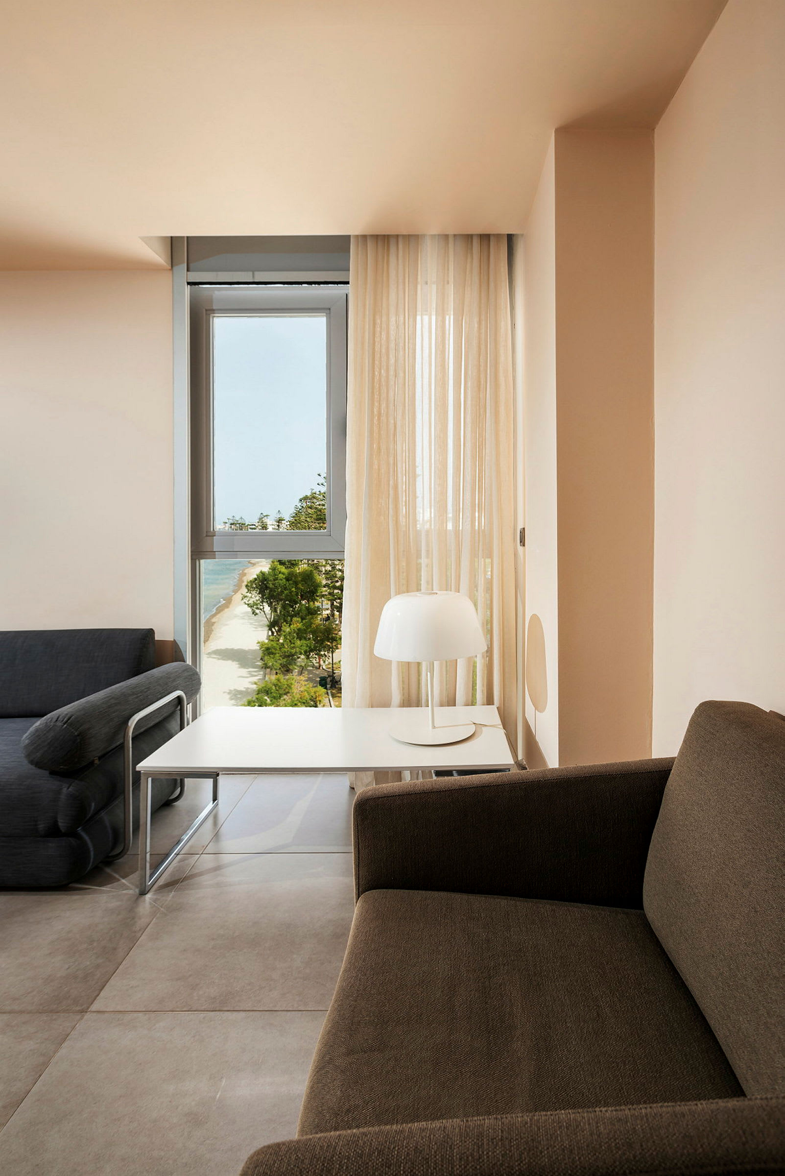 Et rom med en sofa, et bord og en lampe, med utsikt over en strand gjennom et vindu. (AI-generert tekst)