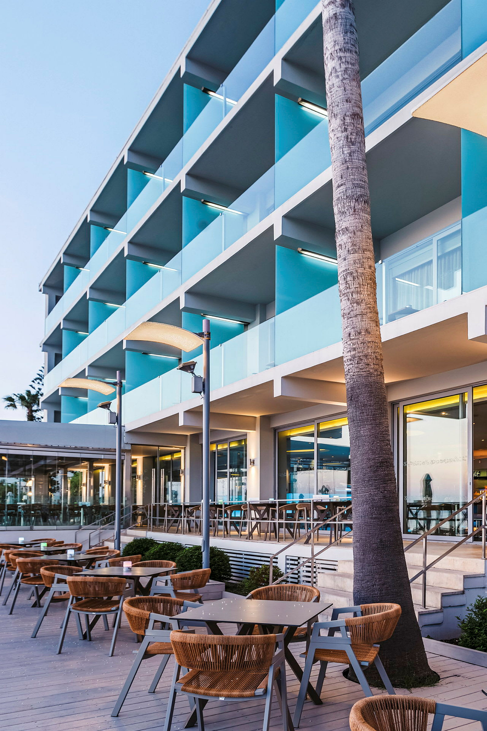 Bilde av en moderne hotellfasade med balkonger og uteservering ved solnedgang. (AI-generert tekst)