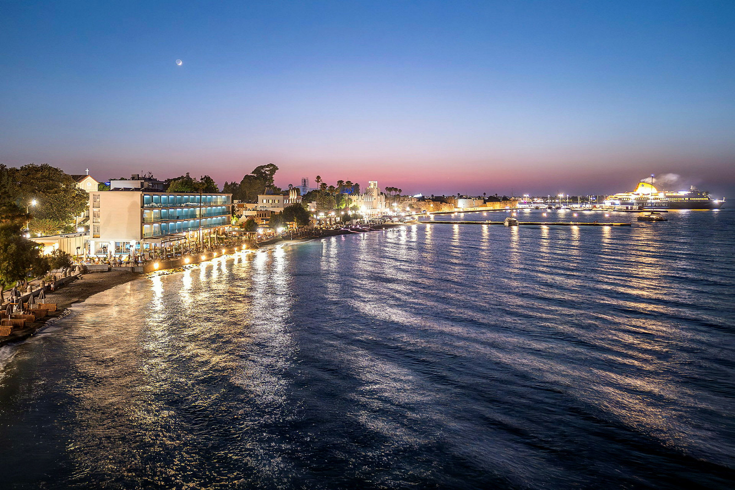 Utsikt over en strandpromenade med bygninger og et ferjeskip ved solnedgang. (AI-generert tekst)