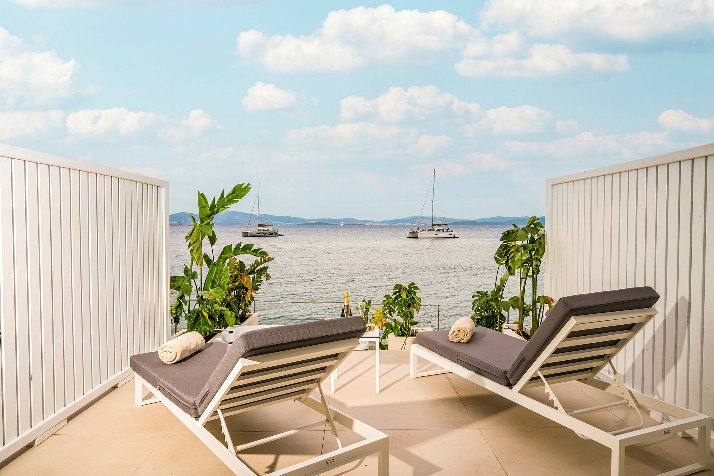 To solsenger med grå puter og håndklær plassert på en terrasse med utsikt over havet og seilbåter. (AI-generert tekst)