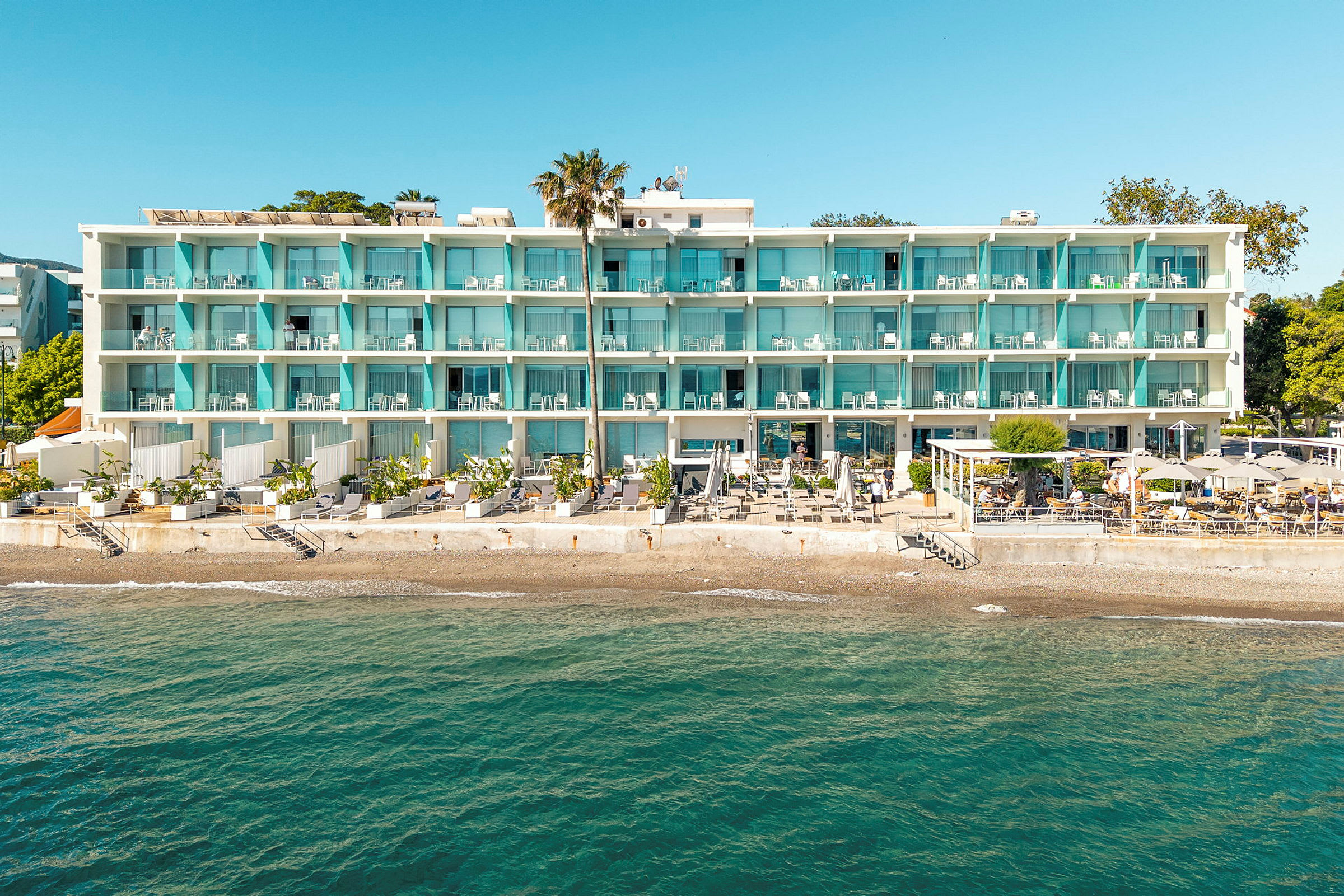 Et hotell ved stranden med en hvit fasade og blå balkonger. Det er en terrasse med parasoller og stoler nedenfor, og havet vises i forgrunnen. (AI-generert tekst)
