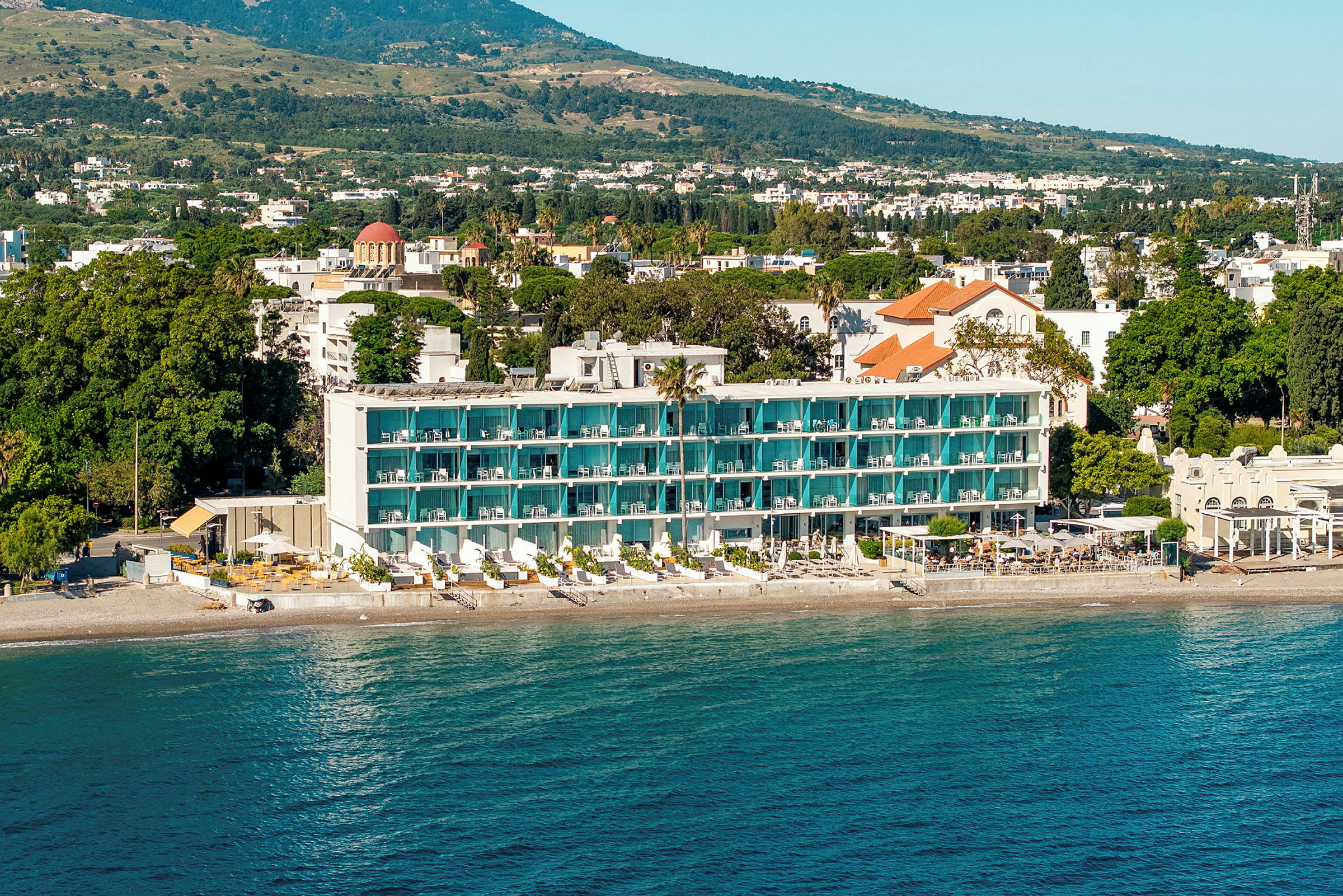 En hotellbygning med balkonger og en strand foran, omgitt av grønne trær og et landskap i bakgrunnen. (AI-generert tekst)