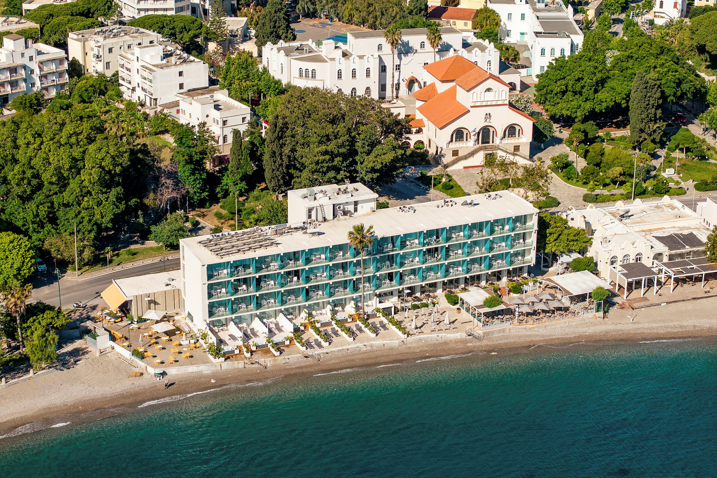 Flyfoto av en hotellbygning ved en strand, omgitt av grøntområder og andre bygninger. (AI-generert tekst)