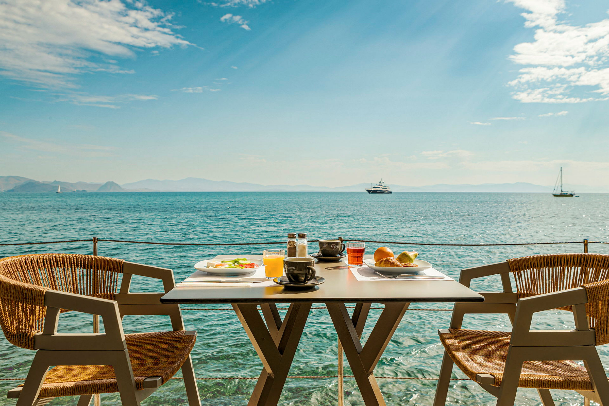 Et dekket bord med frokost står ved havet med båter i bakgrunnen. (AI-generert tekst)