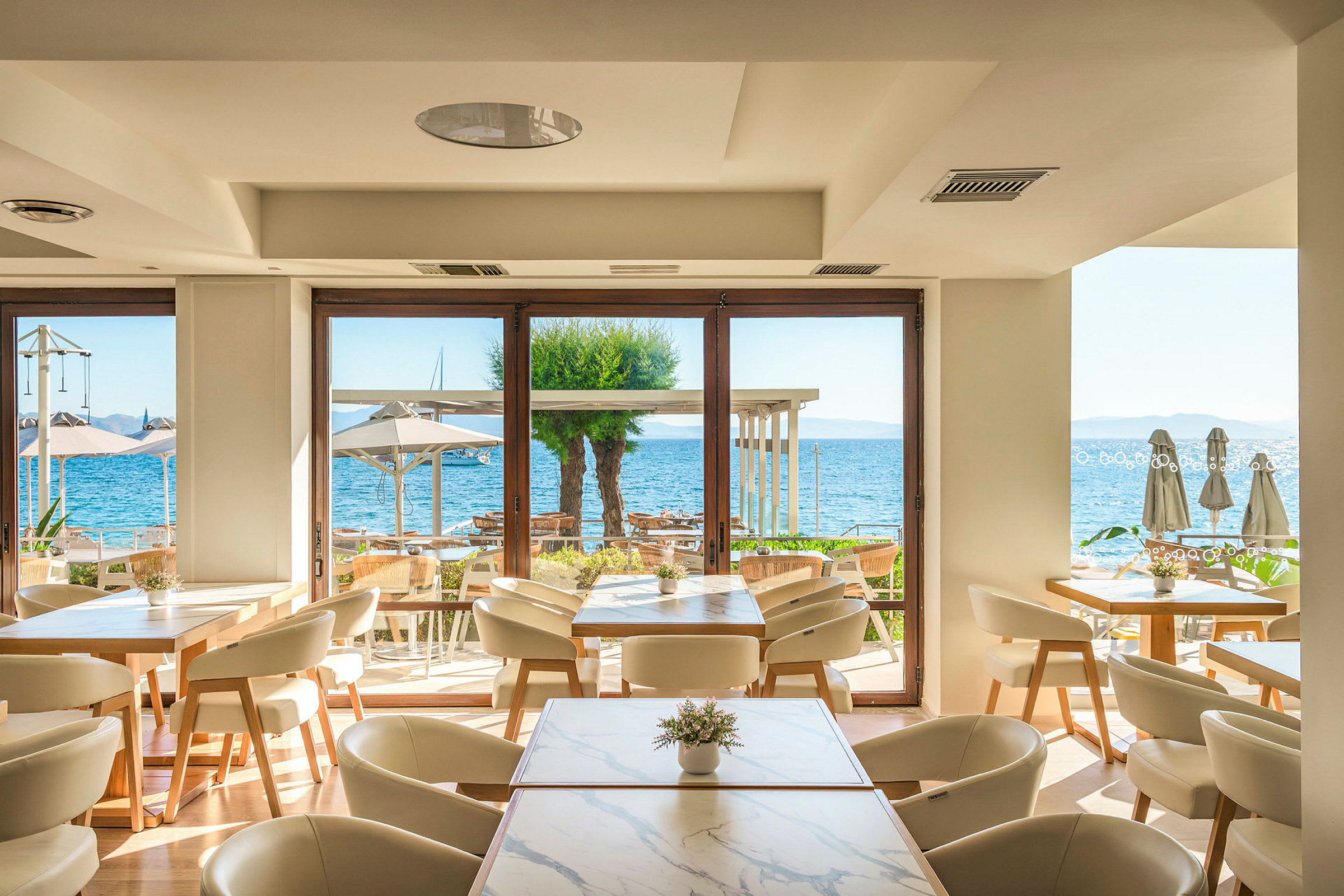 En lys og moderne restaurant med utsikt over havet gjennom store vinduer. Bord med stoler står inne, og utenfor sees parasoller og vann. (AI-generert tekst)