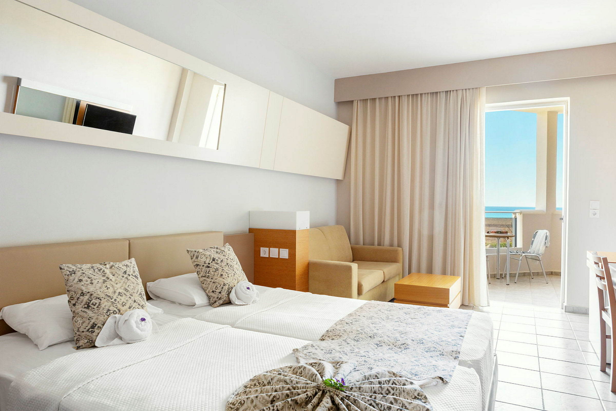 Et hotellrom med to enkeltsenger, en sofa, et bord og en balkong med utsikt mot havet. (AI-generert tekst)