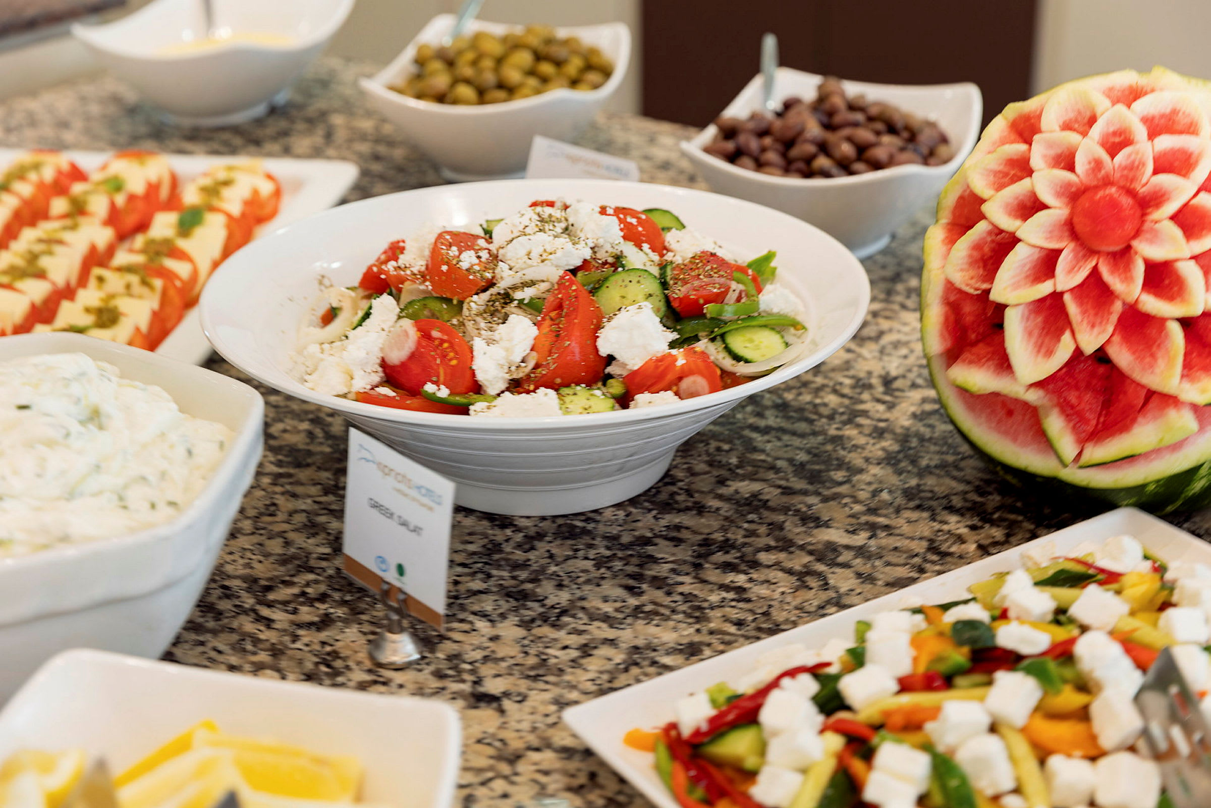 Et buffetbord med salat, grønnsaker, oliven og en dekorativ utskåret vannmelon. (AI-generert tekst)