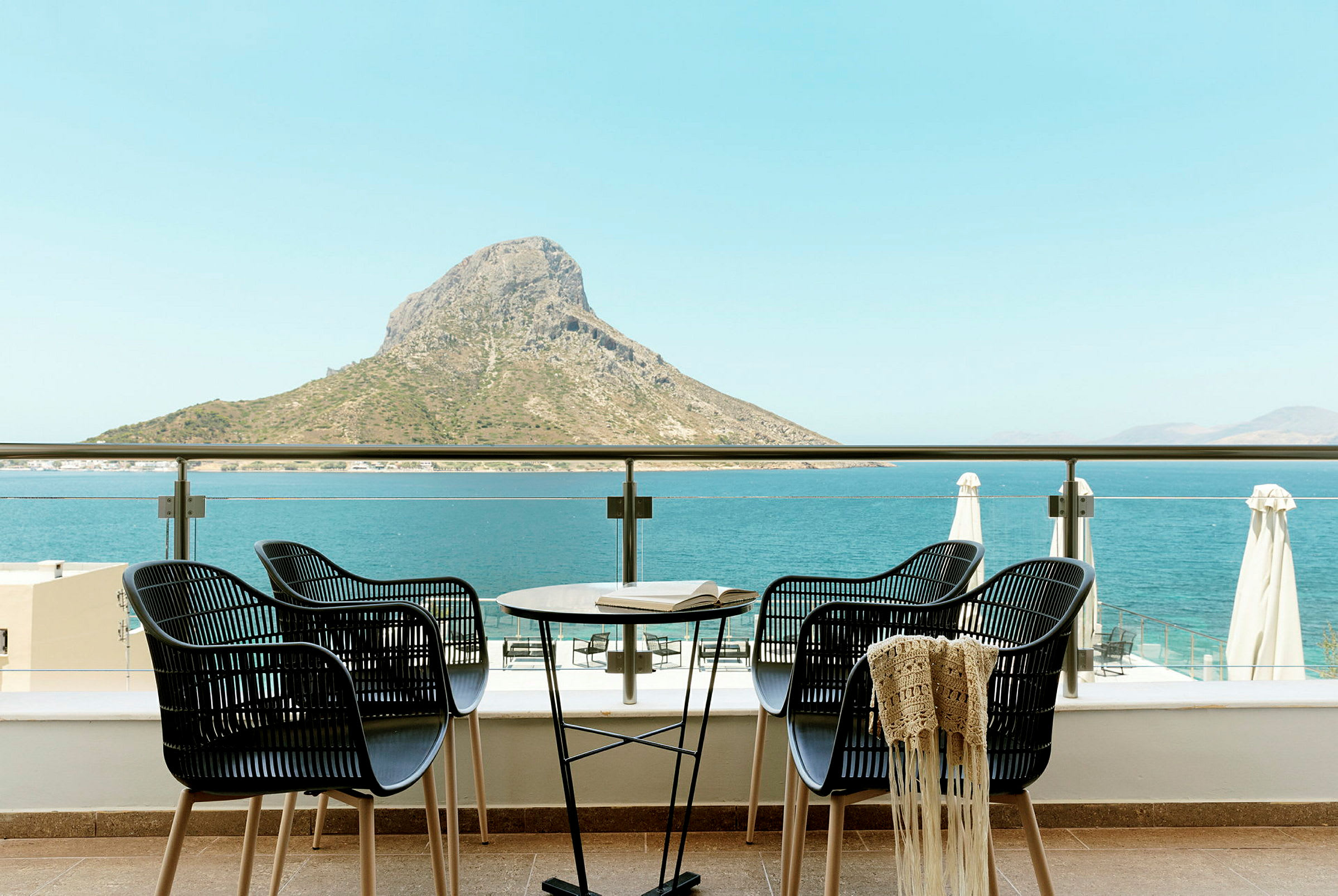 Fire stoler og et bord på en terrasse med utsikt over havet og en fjelltopp. (AI-generert tekst)