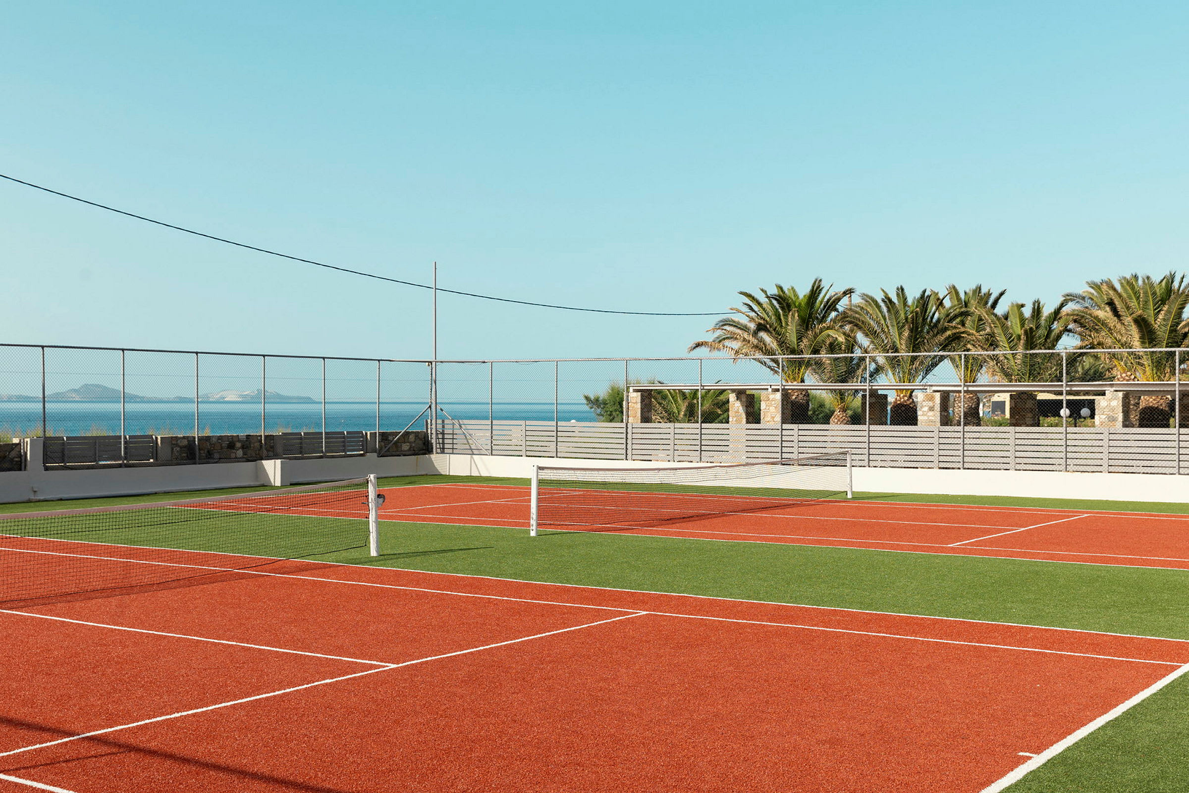 En utendørs tennisbane med rød overflate og grønn omgivelse. I bakgrunnen finnes palmer og hav. (AI-generert tekst)