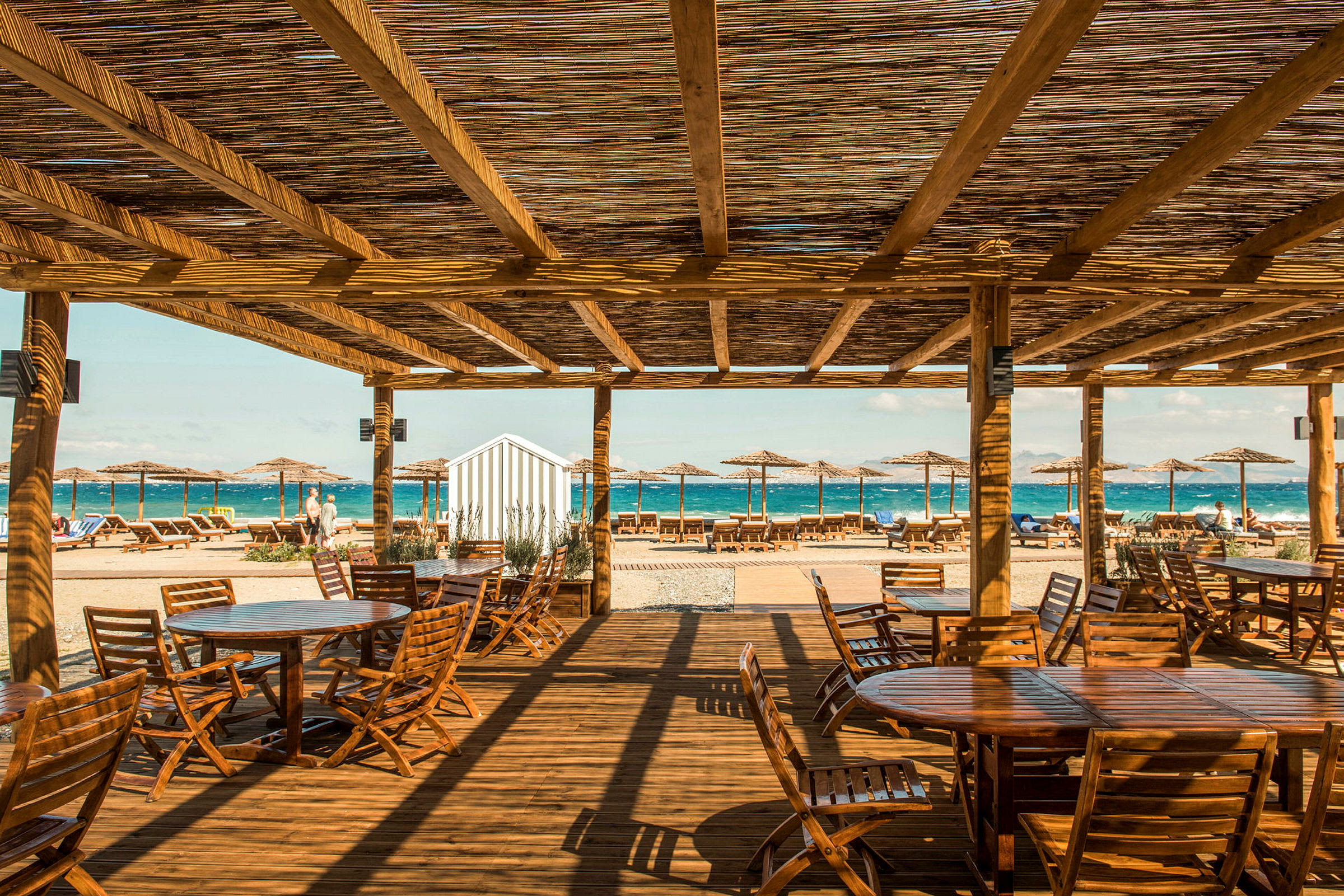 En strandrestaurant med trebord og stoler under et tak av tre. Solsenger og parasoller er plassert ved stranden med havet i bakgrunnen. (AI-generert tekst)