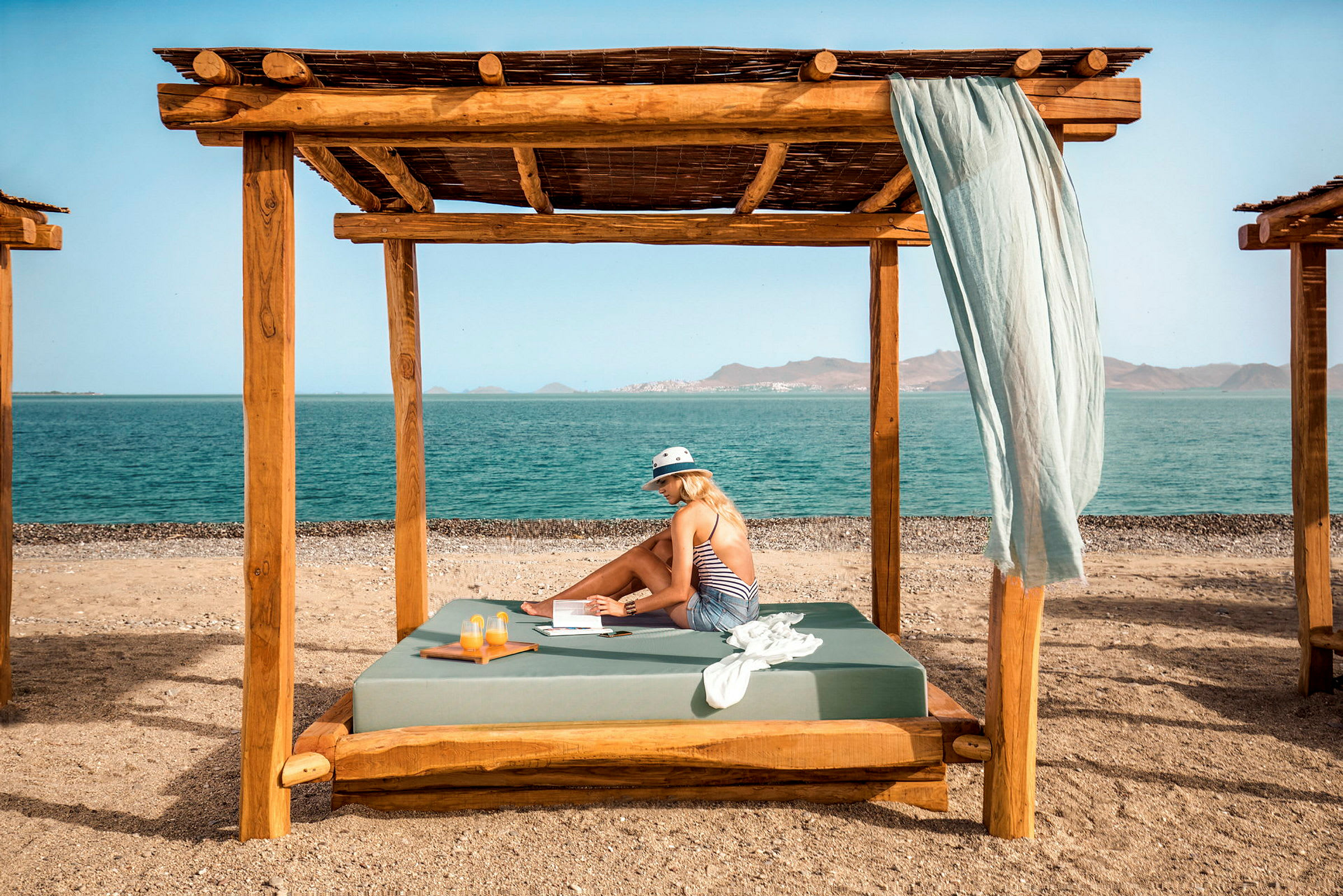 En person sitter under en trepergola på en strand med havet i bakgrunnen. (AI-generert tekst)
