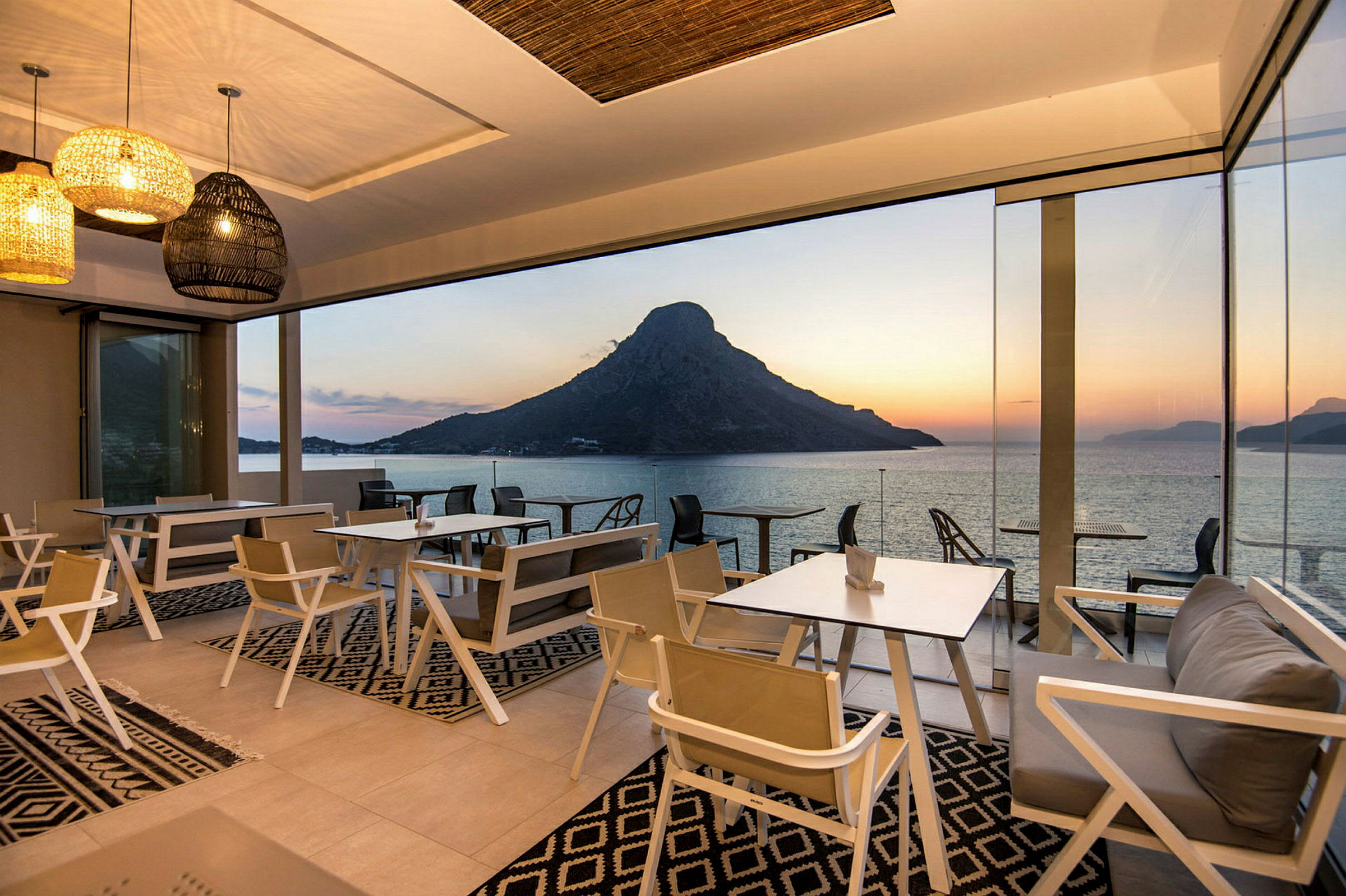 Restaurant med glassveggene og utsikt over havet og fjell ved solnedgang. (AI-generert tekst)