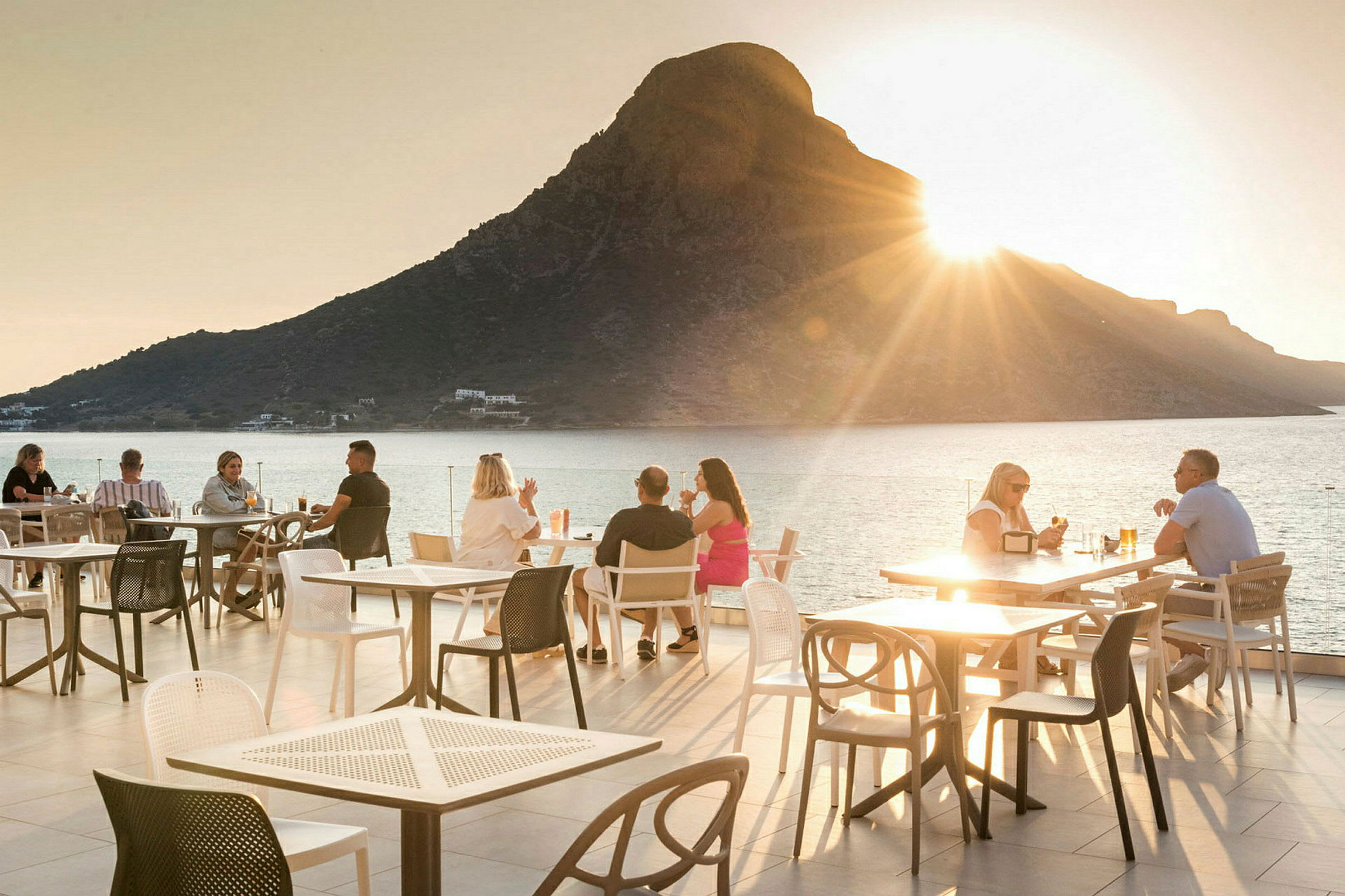 En utendørs terrasse med sitteplasser og et fjell ved havet i bakgrunnen ved solnedgang. (AI-generert tekst)