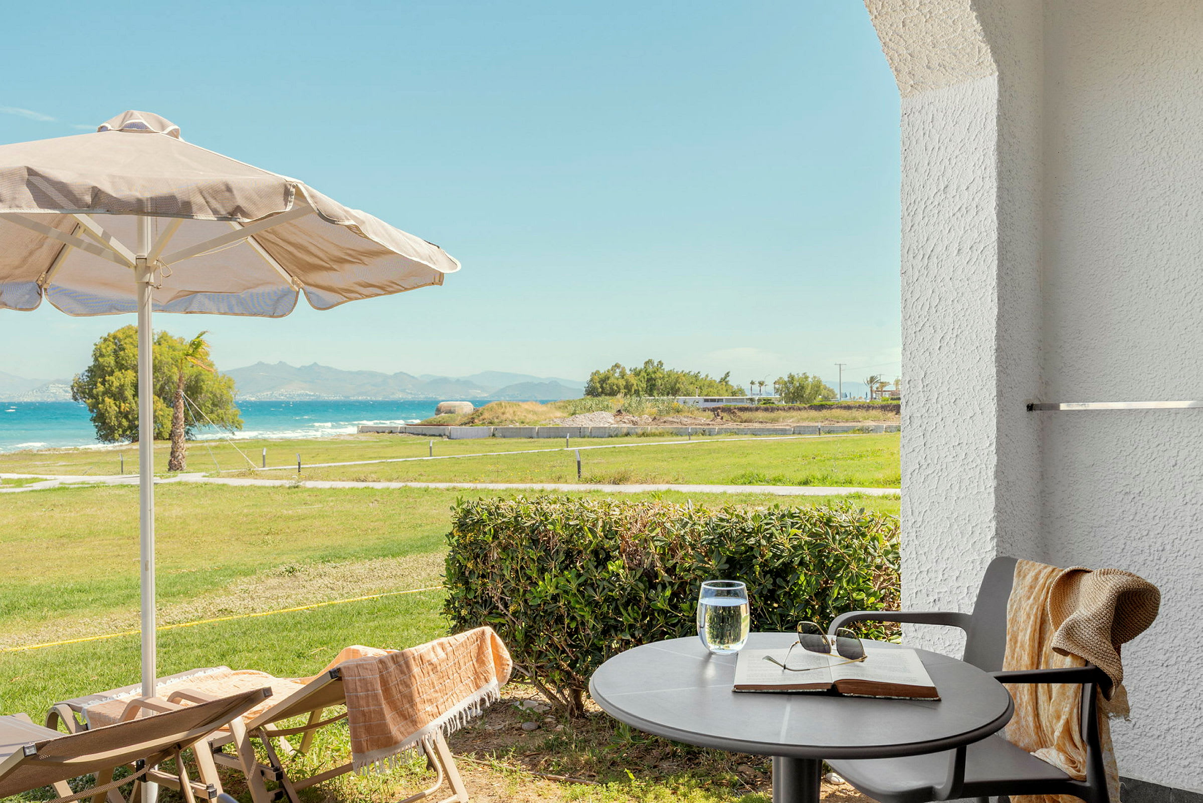 Utsikt fra terrasse med bord, stoler, parasoll og grønt område mot havet. (AI-generert tekst)