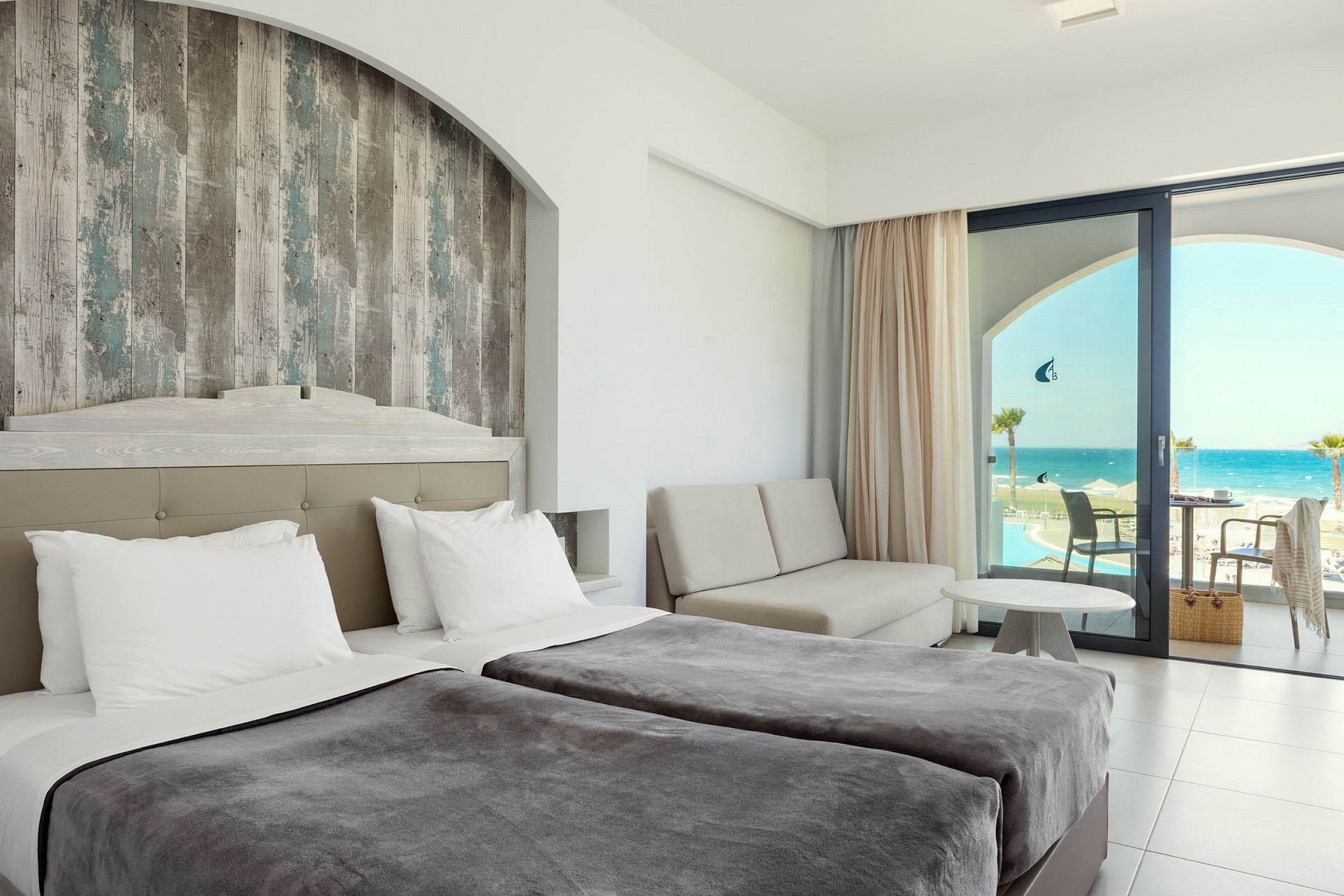 Hotellrom med dobbeltsenger, en sittegruppe og utsikt over havet gjennom en glassdør. (AI-generert tekst)