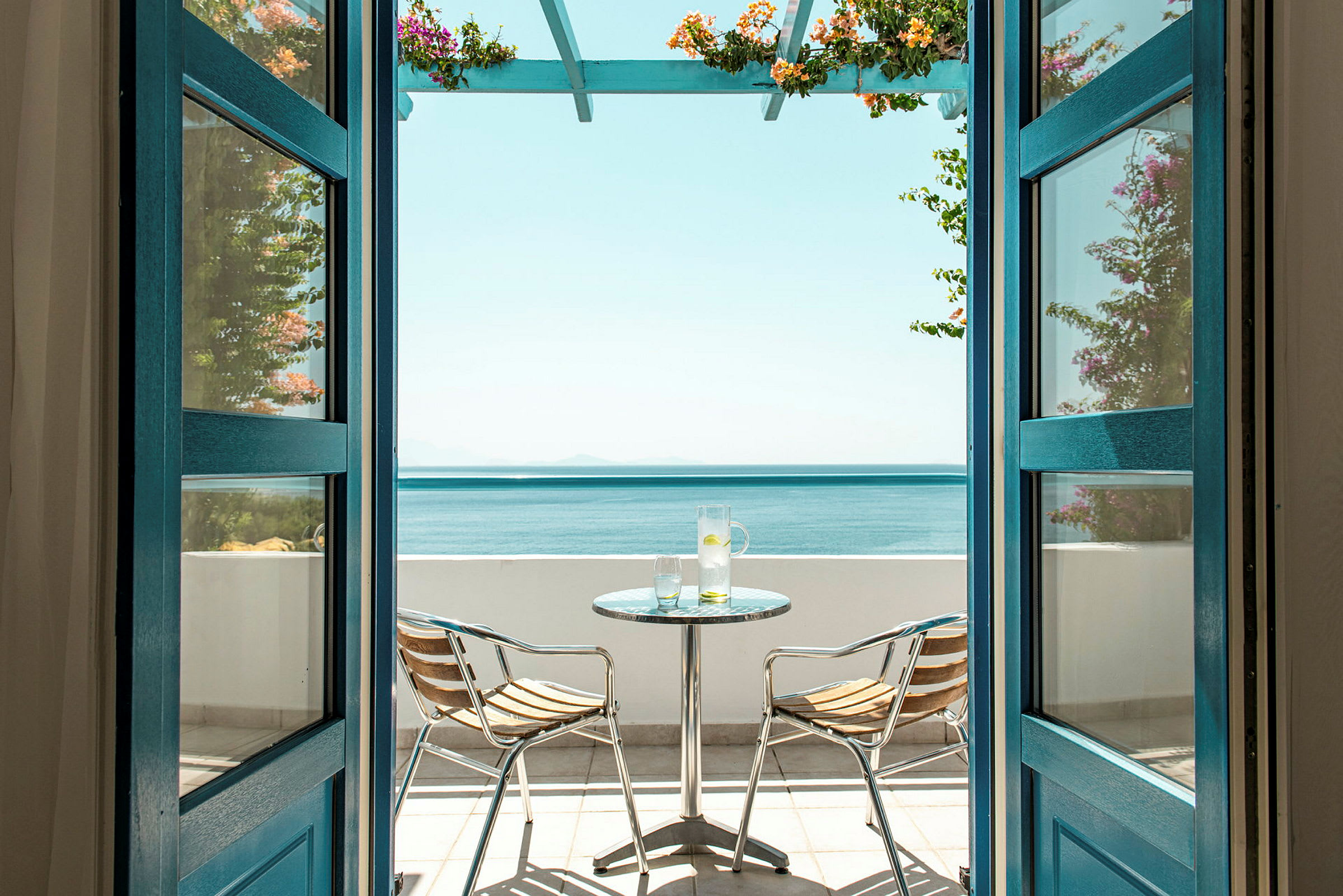 Utsikt gjennom åpne dører mot en terrasse med havutsikt, inkludert et bord og to stoler. (AI-generert tekst)