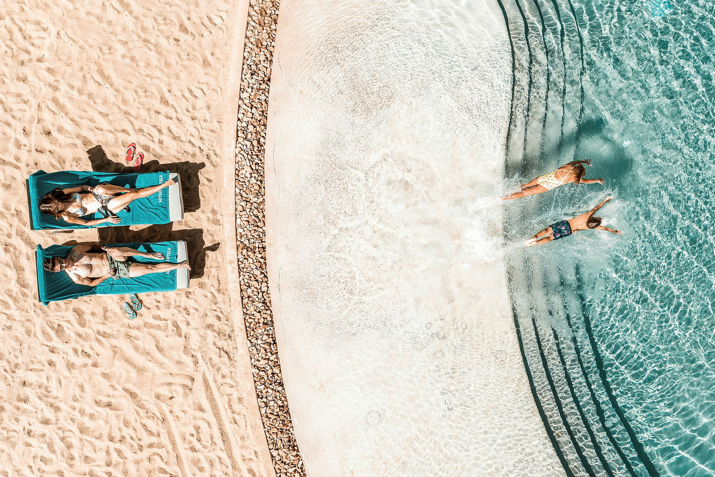 Flyfoto av to personer som ligger på solsenger på en strand ved siden av et klart blått basseng der to andre personer svømmer. (AI-generert tekst)