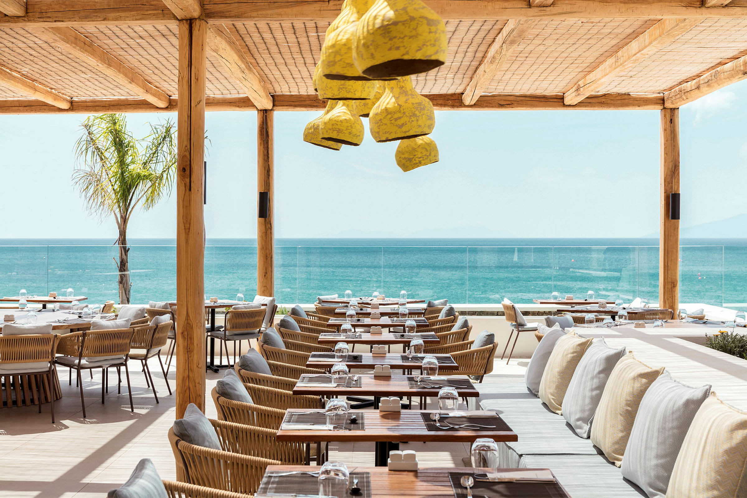 Restaurant med havutsikt, med bord og stoler i tre og rotting samt benker med puter under et beskyttende tak. (AI-generert tekst)