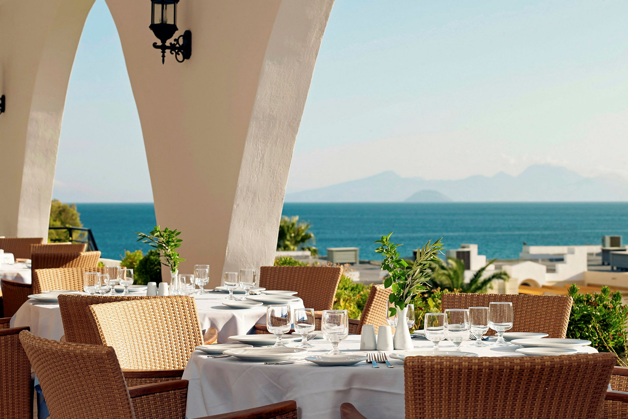 En restaurant med bord dekket med hvite duker, en solfylt utsikt over havet og fjell i bakgrunnen. (AI-generert tekst)