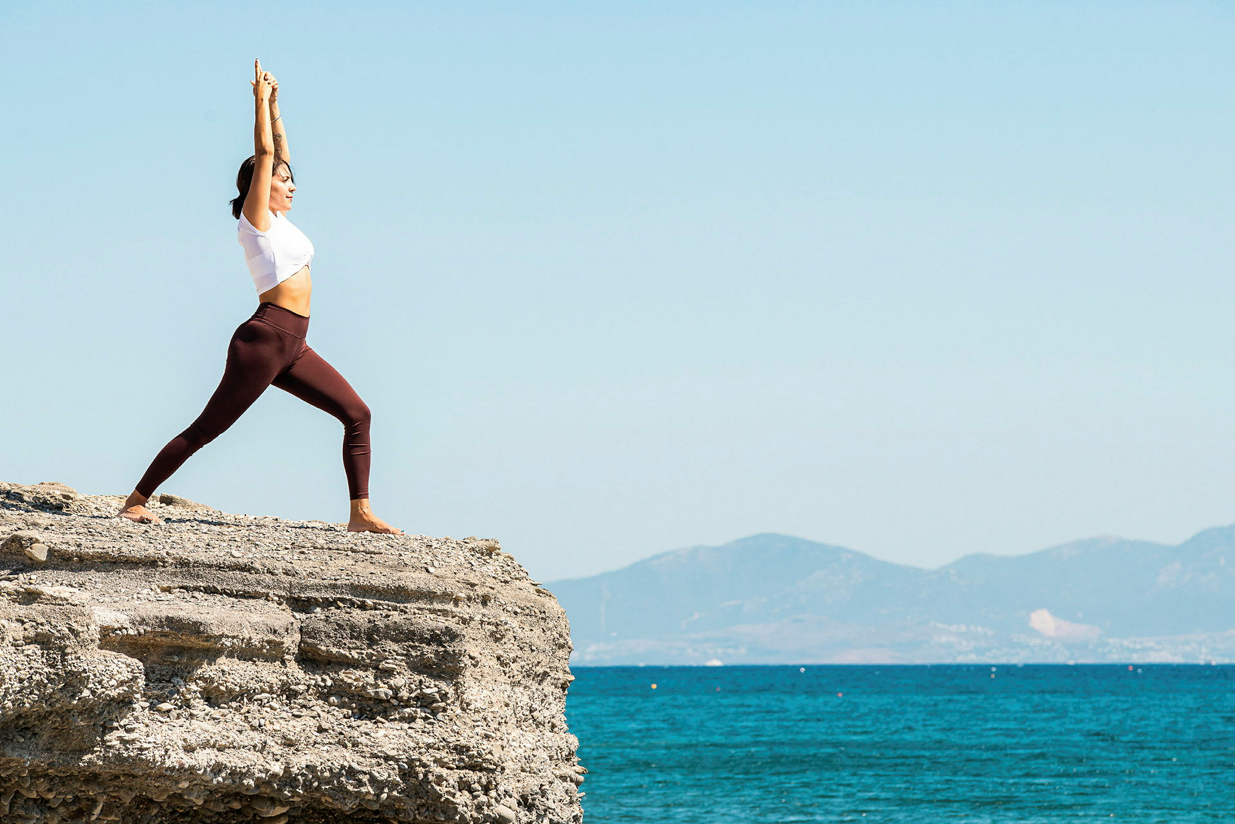 En person driver med yoga på en klippe ved havet med fjell i bakgrunnen. (AI-generert tekst)
