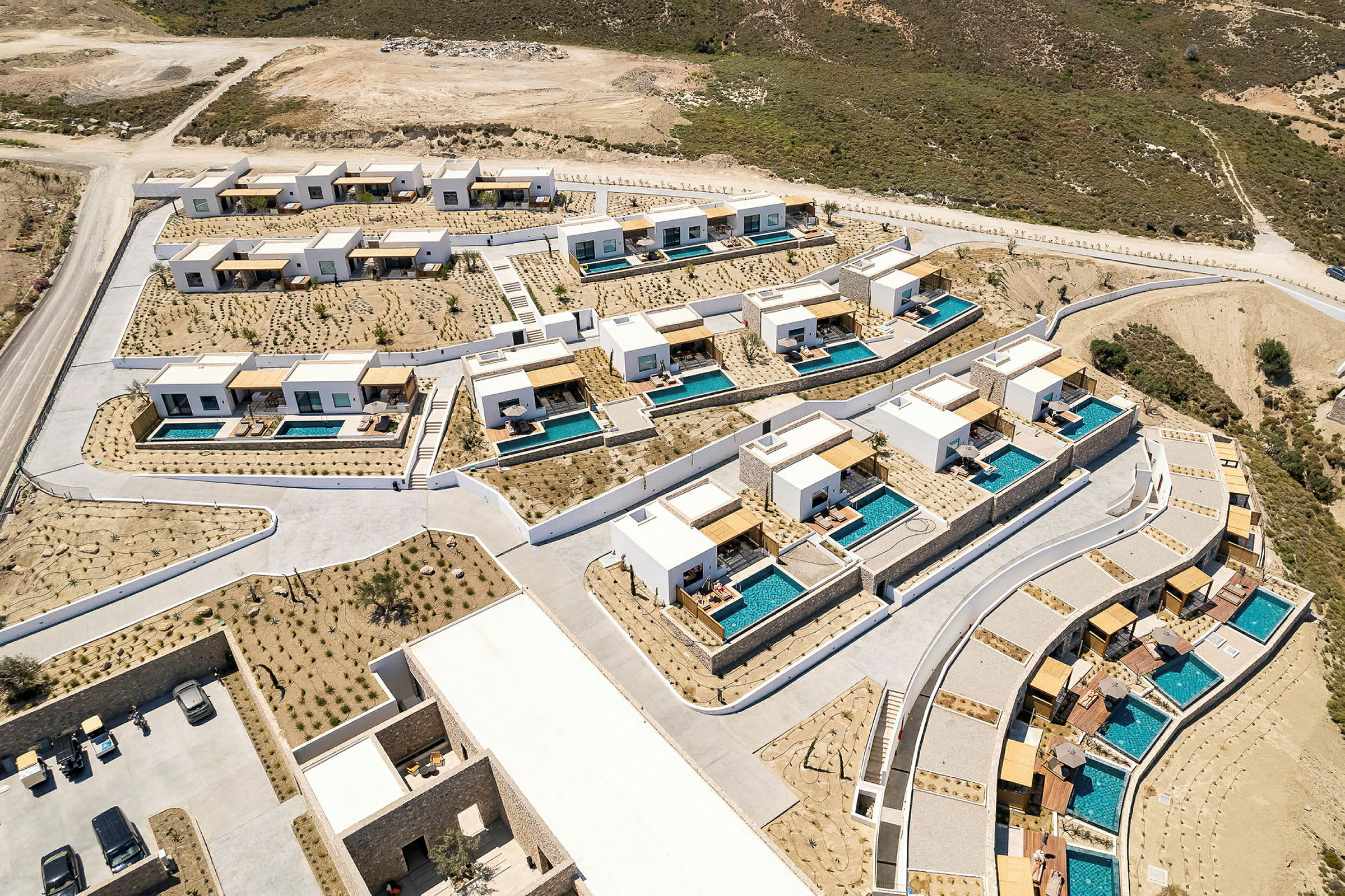 Flyfoto av et ferieanlegg med moderne hus og private bassenger omgitt av tørt landskap. (AI-generert tekst)