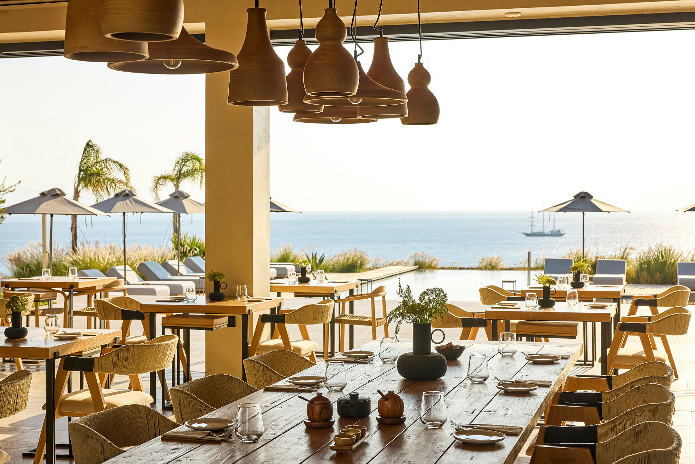 En restaurant med bord og stoler, med utsikt over et utendørs basseng, havet og parasoller. (AI-generert tekst)