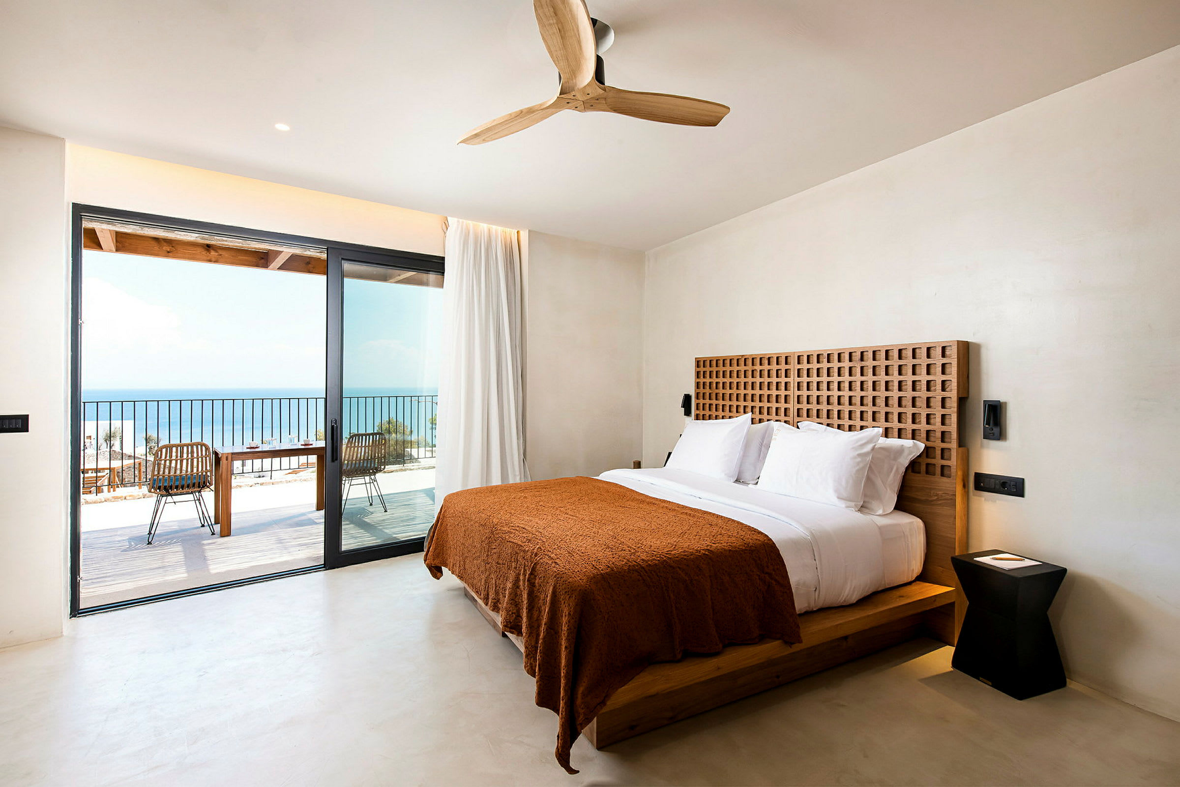 Et moderne hotellrom med en dobbeltseng med hvitt sengetøy og et brunt teppe. Rommet har en stor glassdør som fører til en balkong med utsikt over havet. (AI-generert tekst)