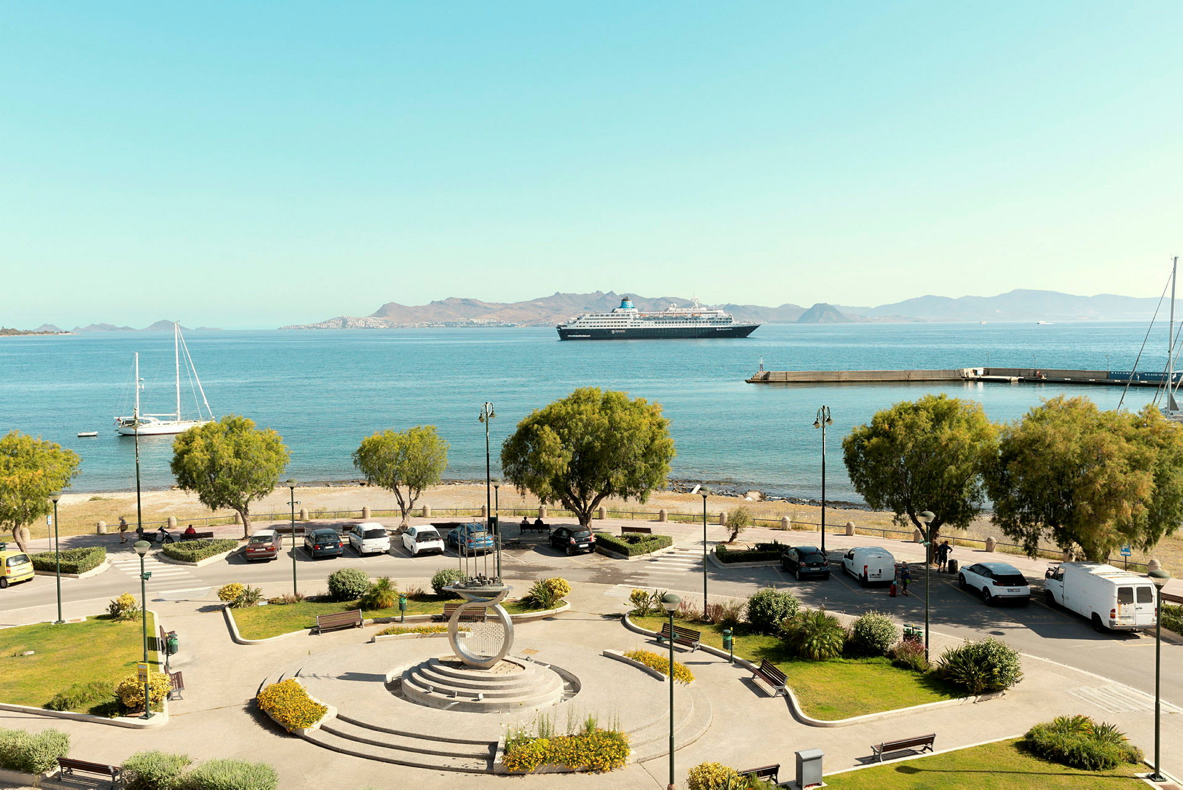 Utsikt over en havn med parkeringsplass og trær i forgrunnen, båter og et cruiseskip på det blå havet, samt fjell i bakgrunnen. (AI-generert tekst)