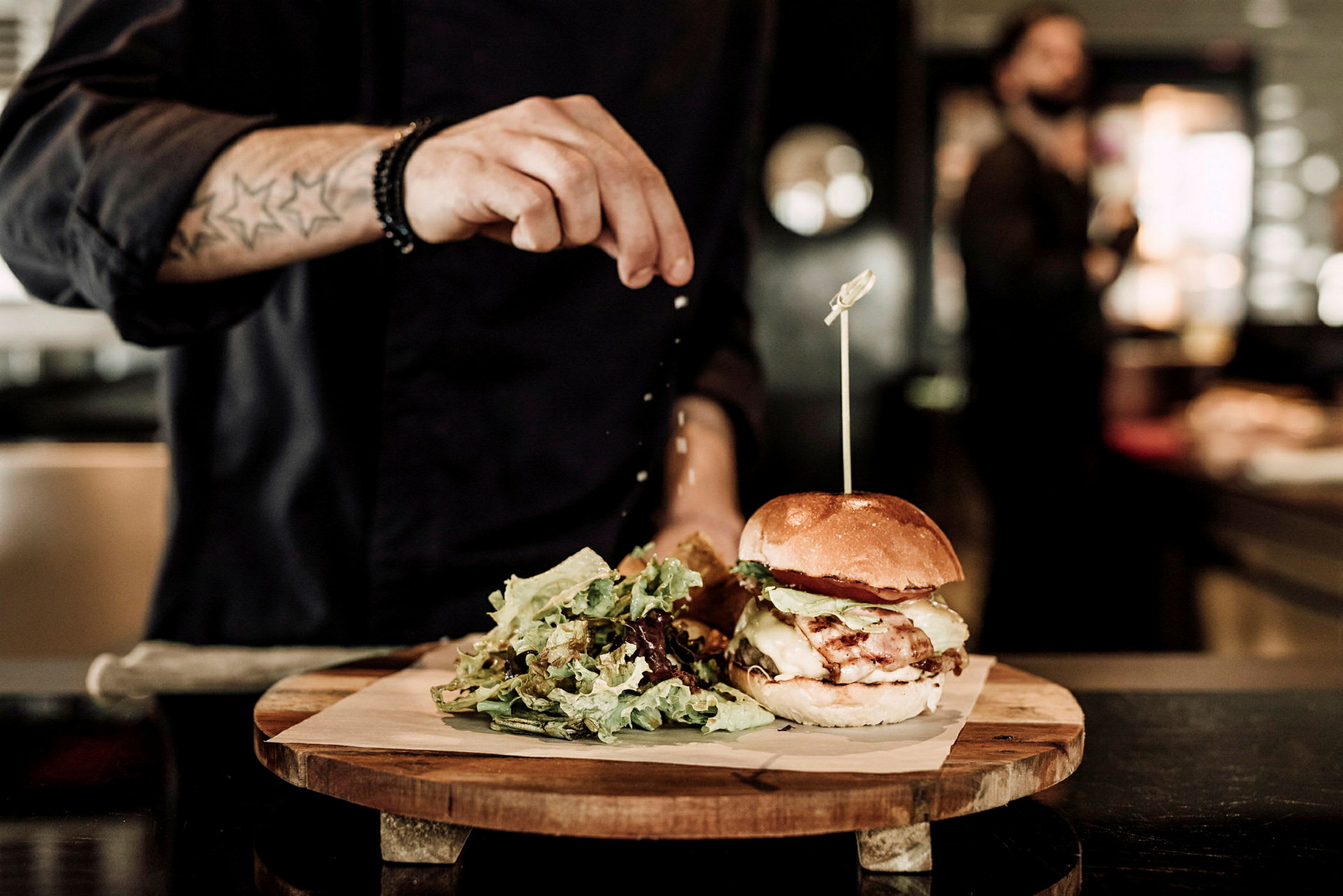 En person krydrer en hamburger som er plassert på en treplate sammen med en porsjon salat. (AI-generert tekst)