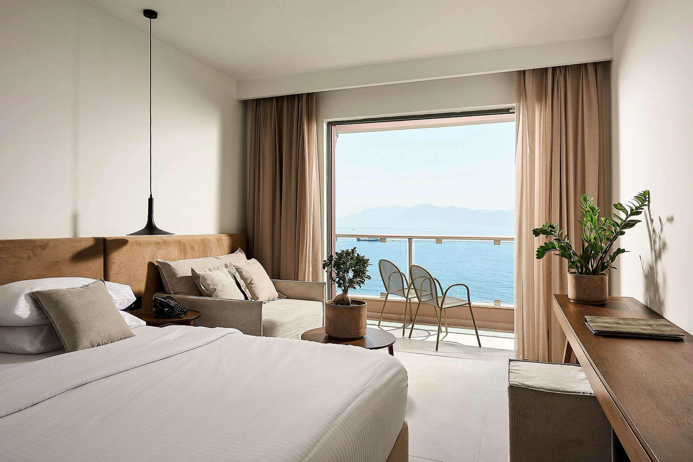 Et hotellrom med en seng, sofa og utsikt over havet fra en balkong. (AI-generert tekst)