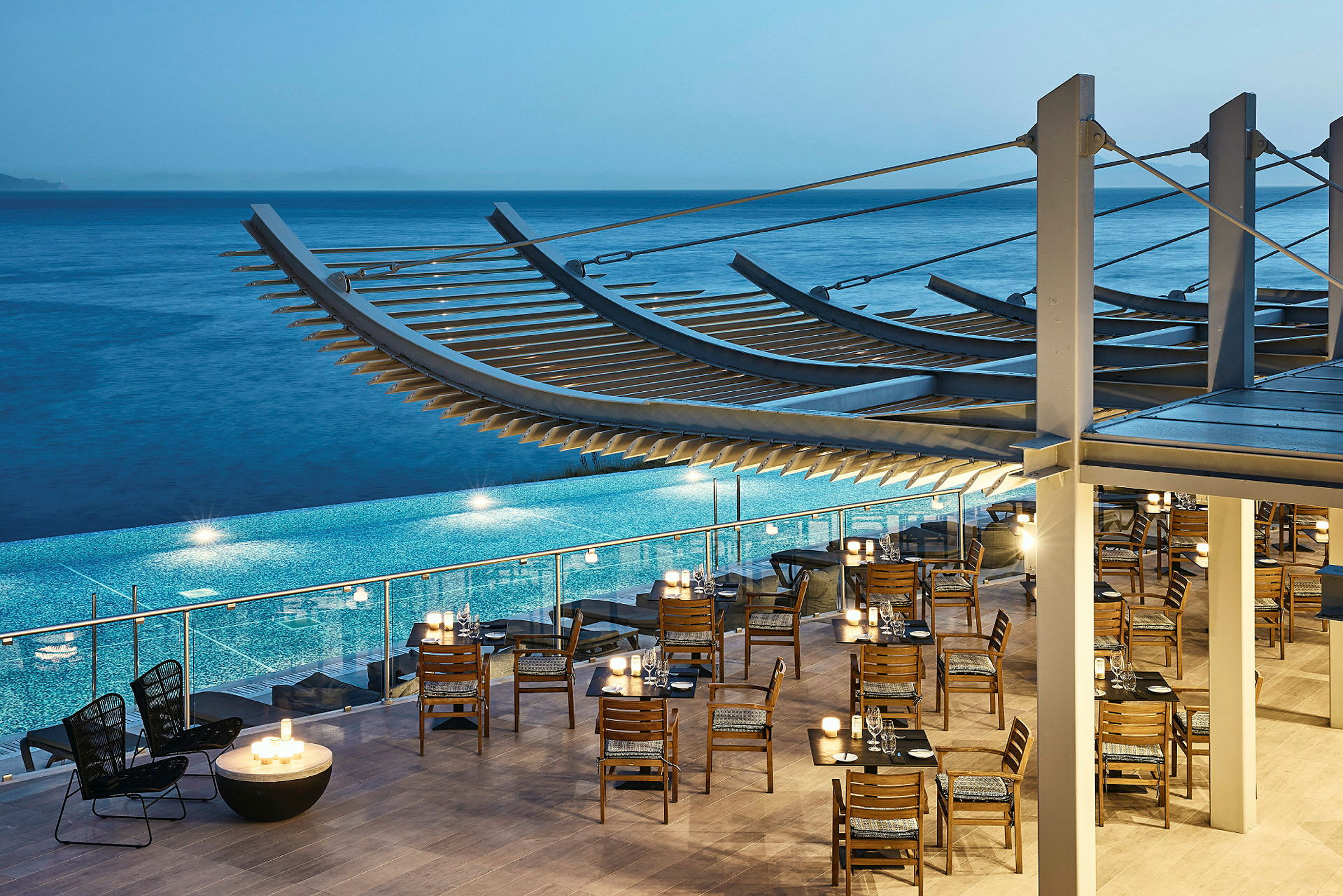 Restaurant med trestoler og bord ved et basseng, havet i bakgrunnen. (AI-generert tekst)
