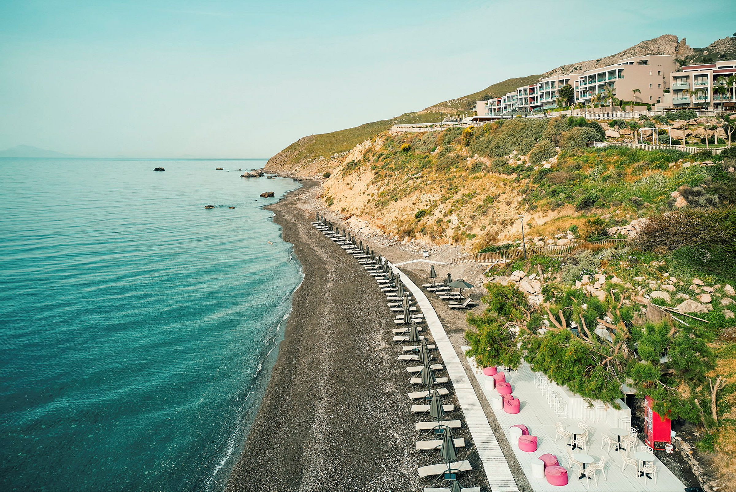 Strand med parasoller og solsenger langs en sti, bratte skråninger i bakgrunnen. (AI-generert tekst)