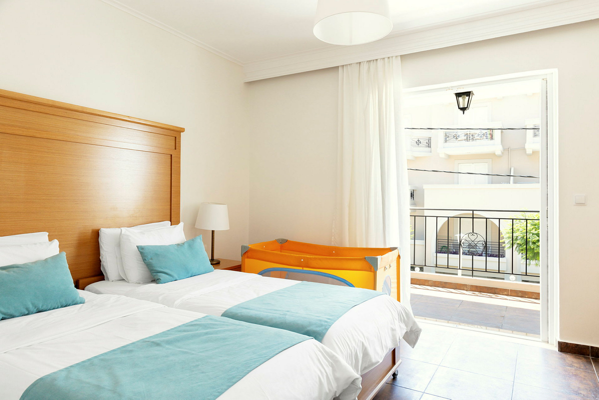 Hotellrom med to enkeltsenger, lyse møbler og en oransje barneseng. (AI-generert tekst)
