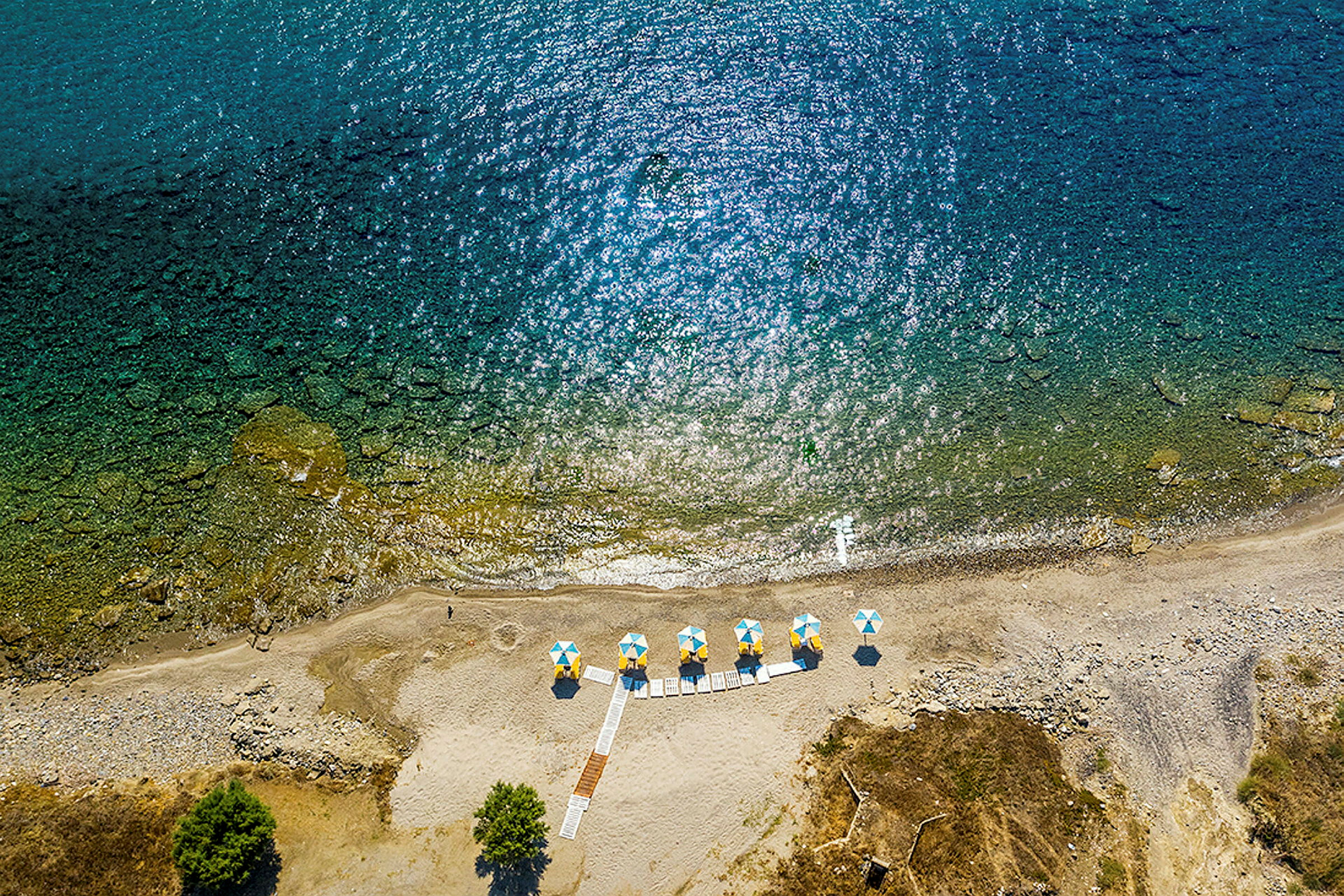 Luftfoto av en strand med parasoller i gule og blå farger samt gangveier. (AI-generert tekst)