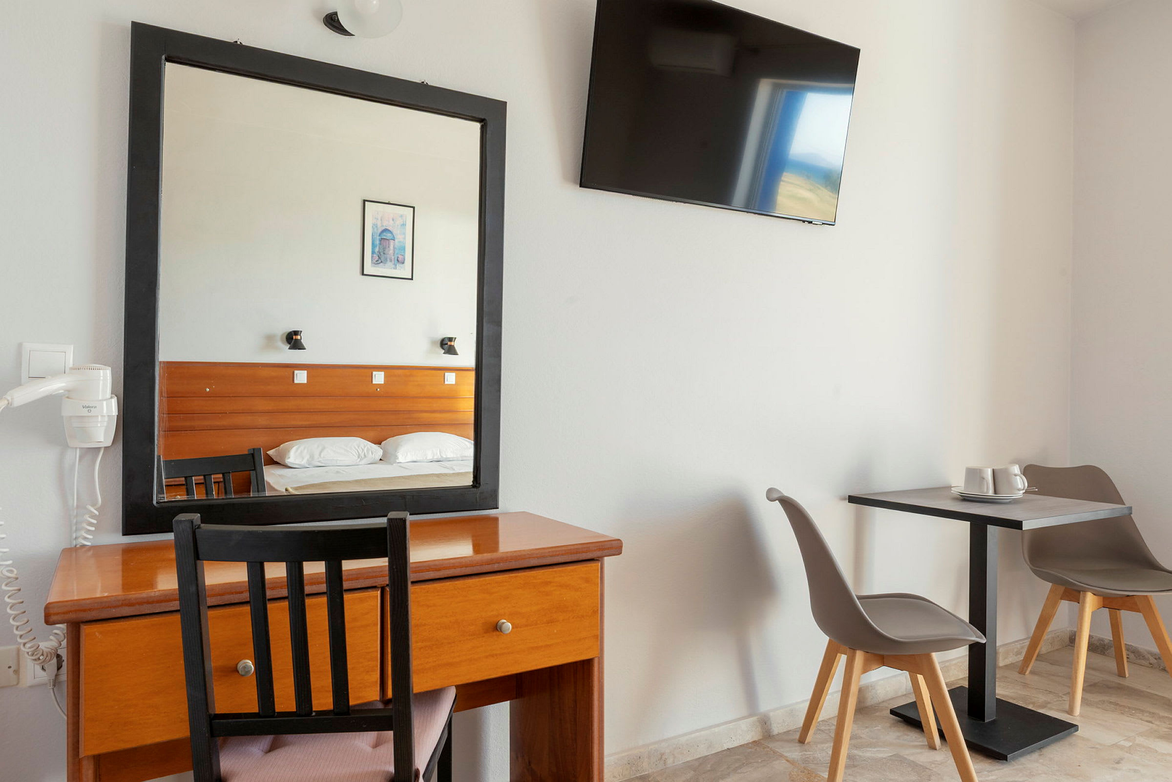 Hotellrom med skrivebord, speil og TV på veggen. Til høyre to stoler ved et bord. (AI-generert tekst)