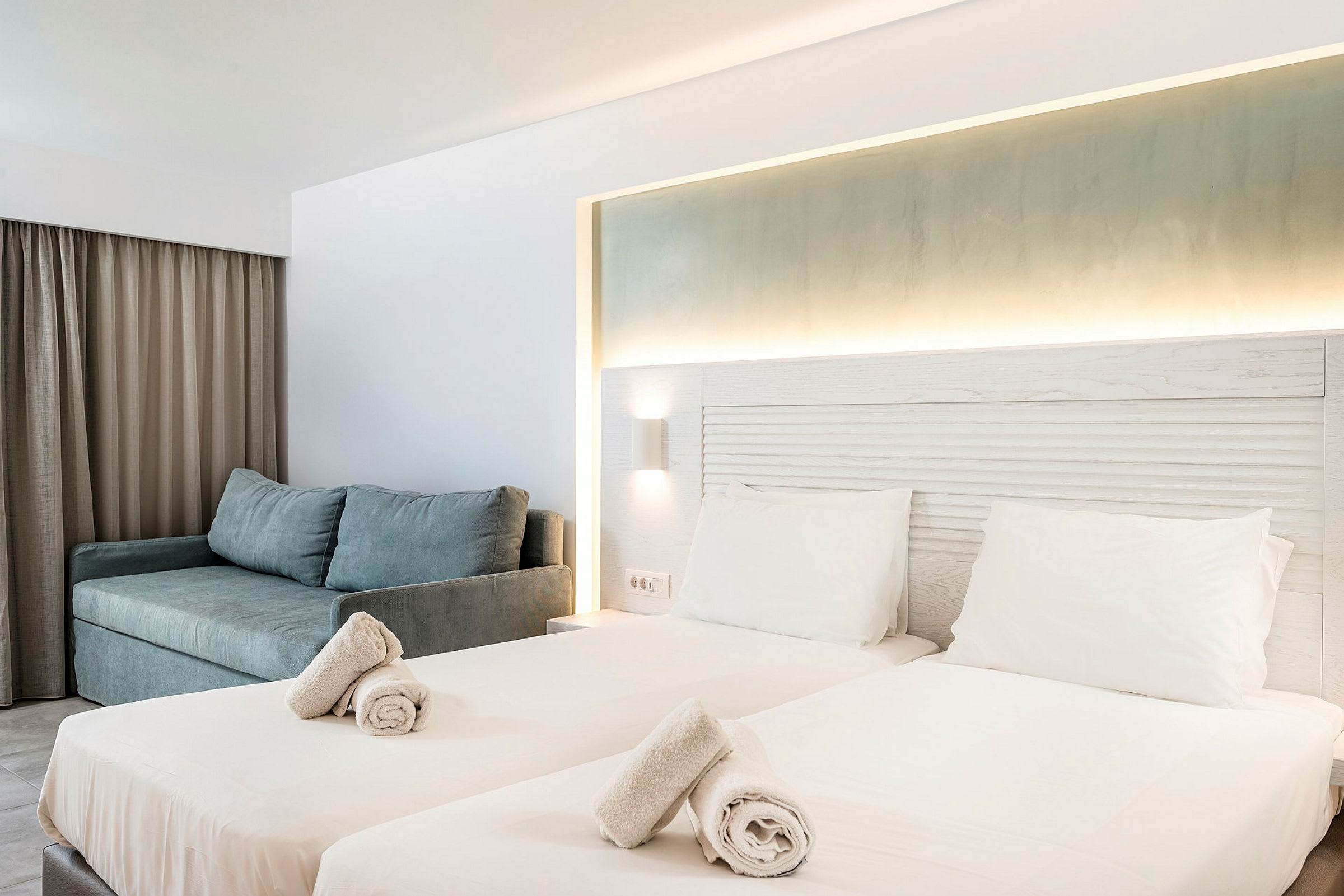 Hotellrom med to enkelsenger, opplyst sengegavl og en sofa. (AI-generert tekst)