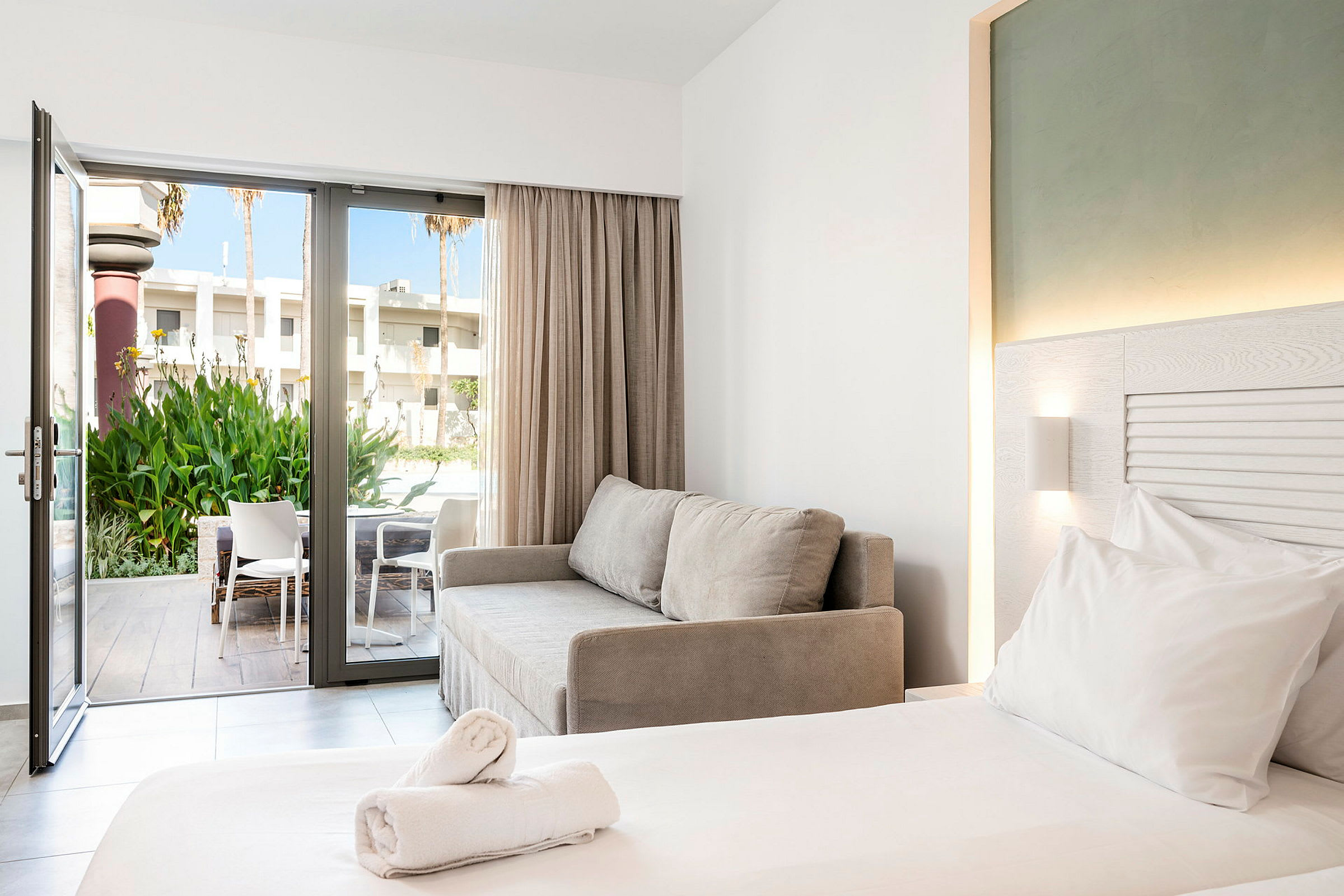 Et moderne hotellrom med en dobbeltseng, en grå sofa og en åpen glassdør som fører til en terrasse med stoler og grøntområder. (AI-generert tekst)