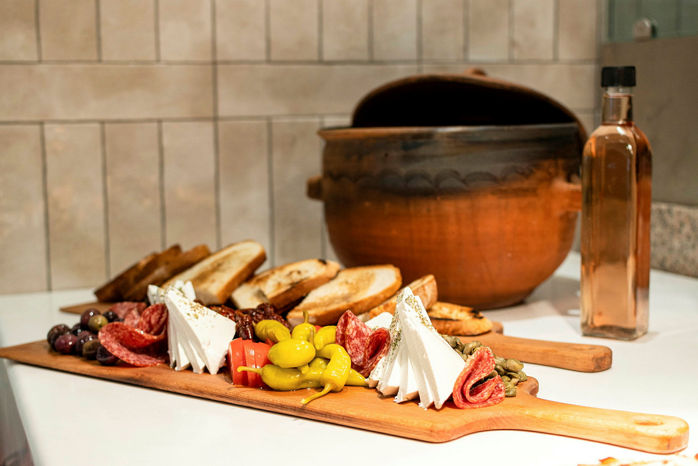 Et skjærebrett med ulike antipasti som ost, salami, oliven og brød, samt en leirgryte og en glassflaske i bakgrunnen. (AI-generert tekst)