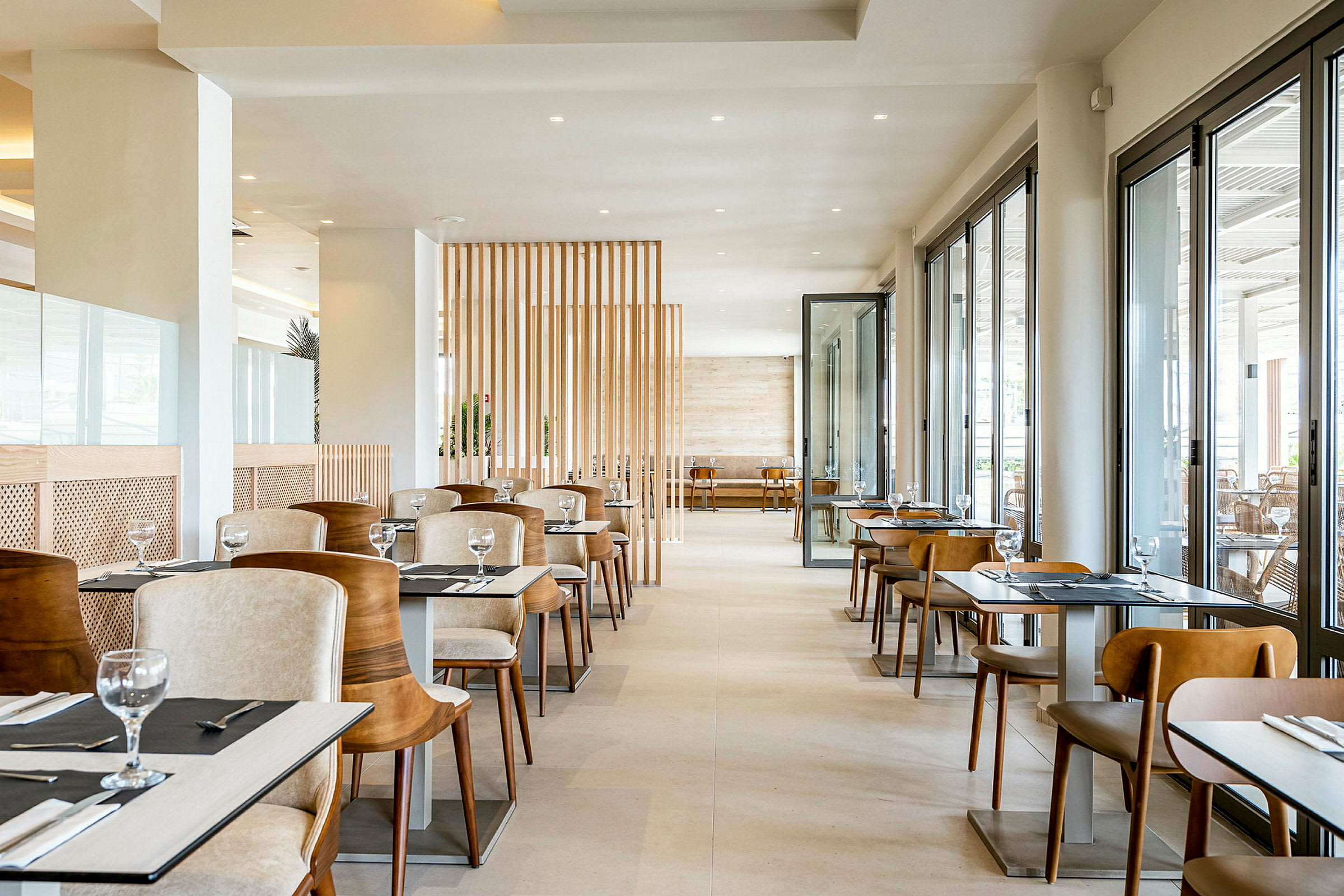 Restaurantinteriør med bord og stoler. Området er lyst og moderne med store vinduer som slipper inn dagslys. (AI-generert tekst)