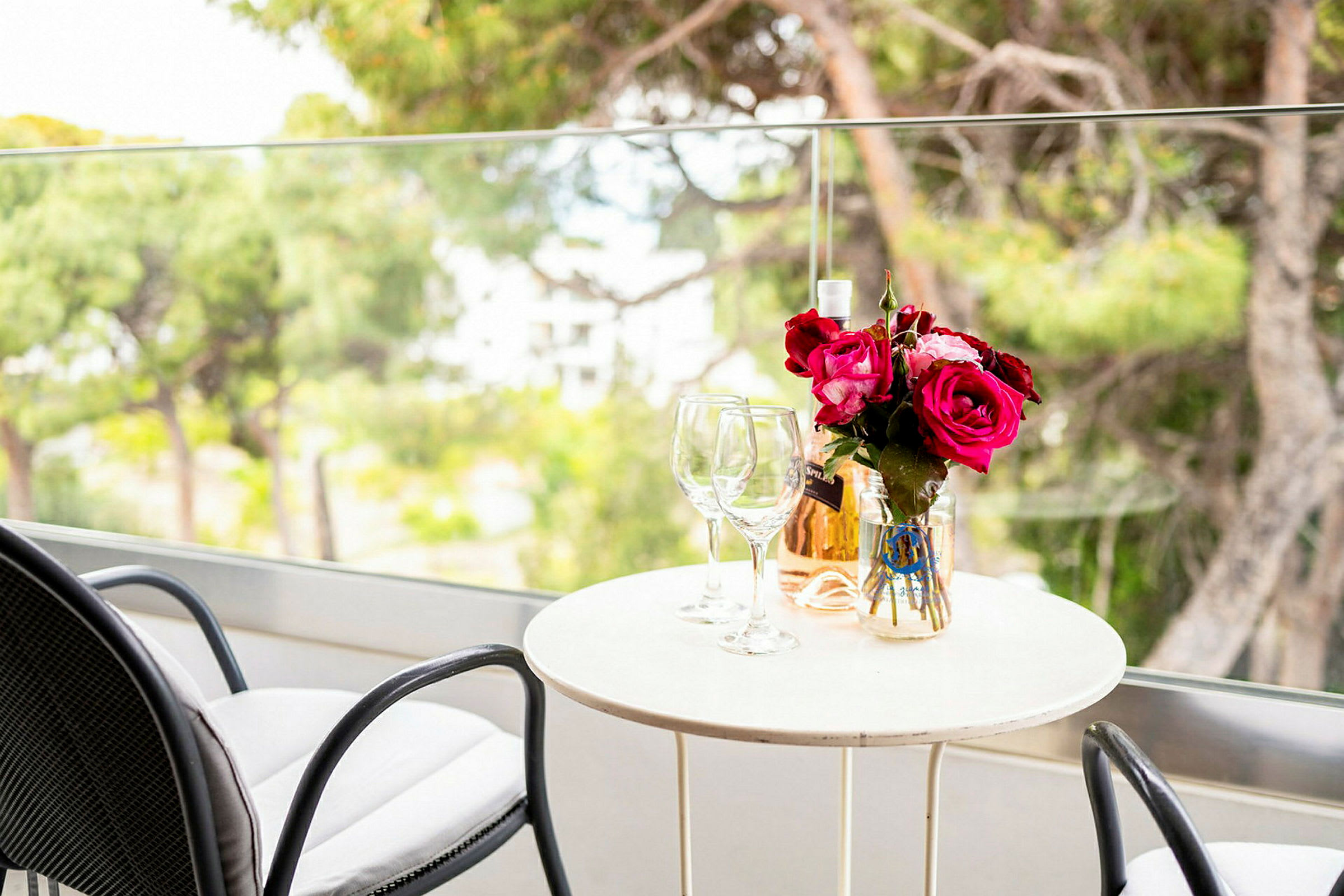 Et bord med to glass, en vase med rosa blomster og en flaske rosévin på en balkong. (AI-generert tekst)