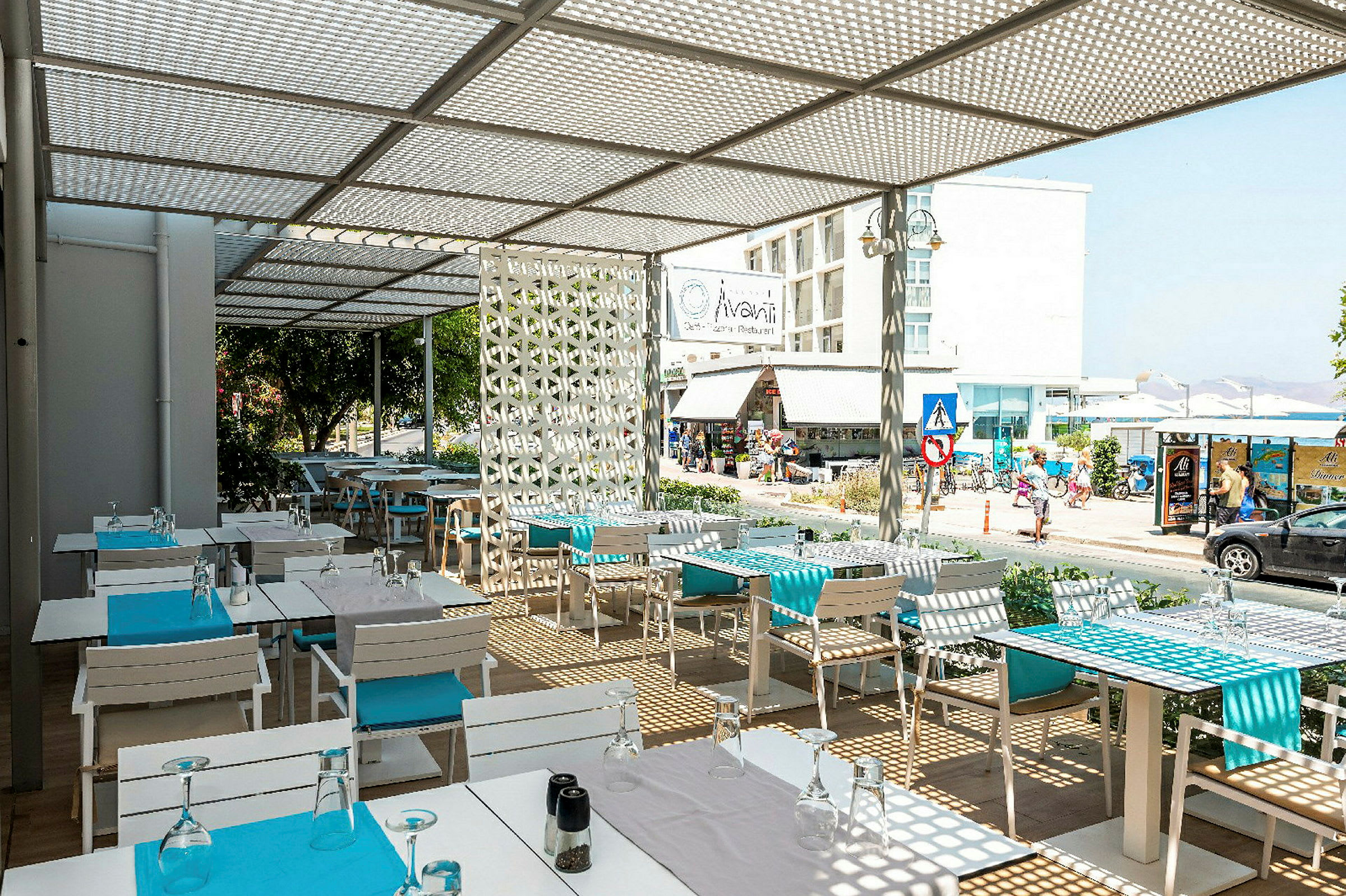 En utendørs restaurant med hvite bord og blå detaljer, med utsikt mot en gate og bygninger. (AI-generert tekst)