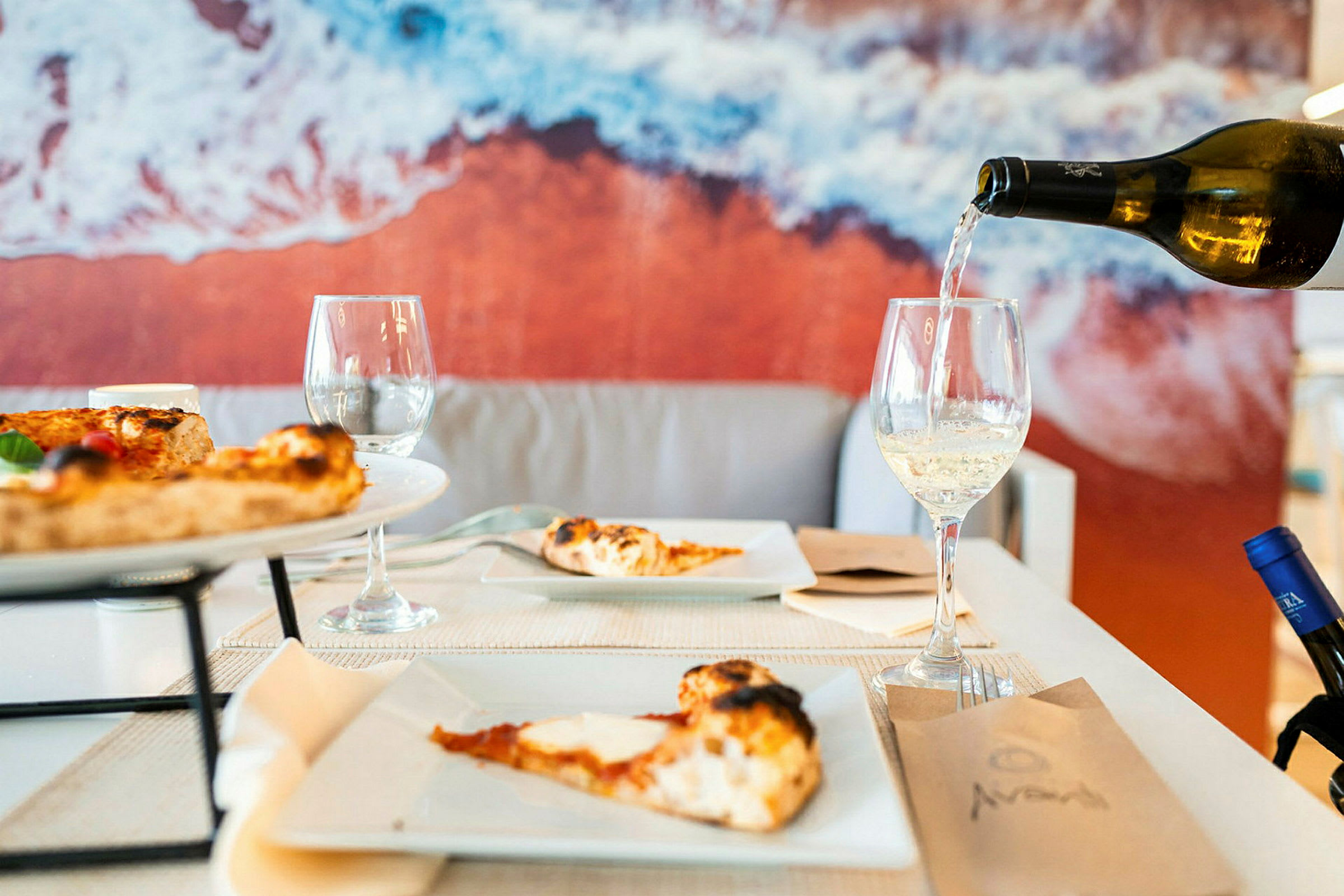 Et bord med pizza og vinglass, bakgrunn med et bilde av stranden. (AI-generert tekst)