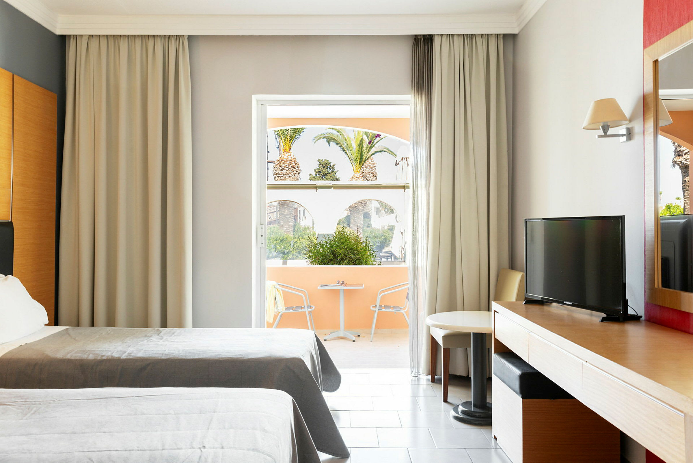 Hotellrom med to senger, skrivebord, TV og en sittegruppe ved et vindu som vender mot en terrasse med utsikt over palmer. (AI-generert tekst)